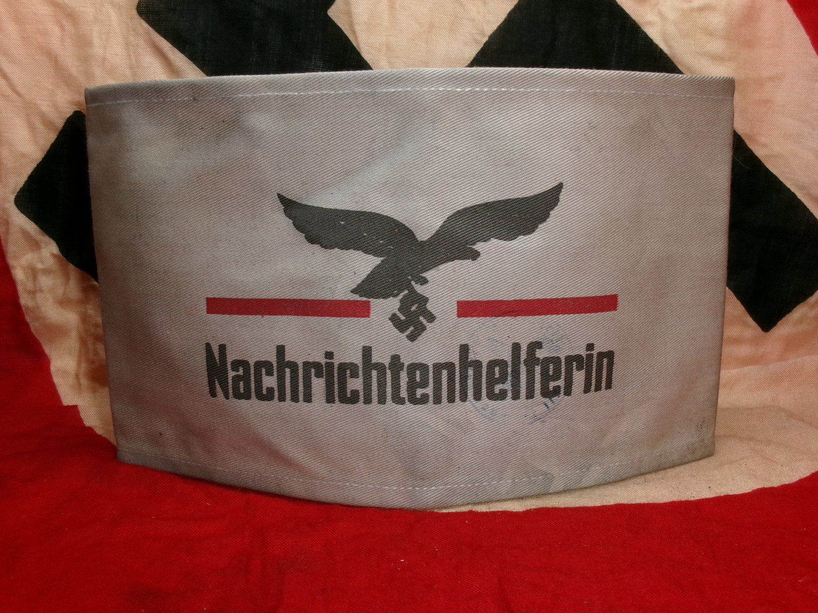 WWII German Luftwaffe Nachrichtenhelferin Armband (Signals Auxiliary Identification): A grey cloth armband worn by female Luftwaffe communications auxiliaries, printed with the word “Nachrichtenhelferin” beneath a Luftwaffe eagle clutching a swastika. Red horizontal bars frame 