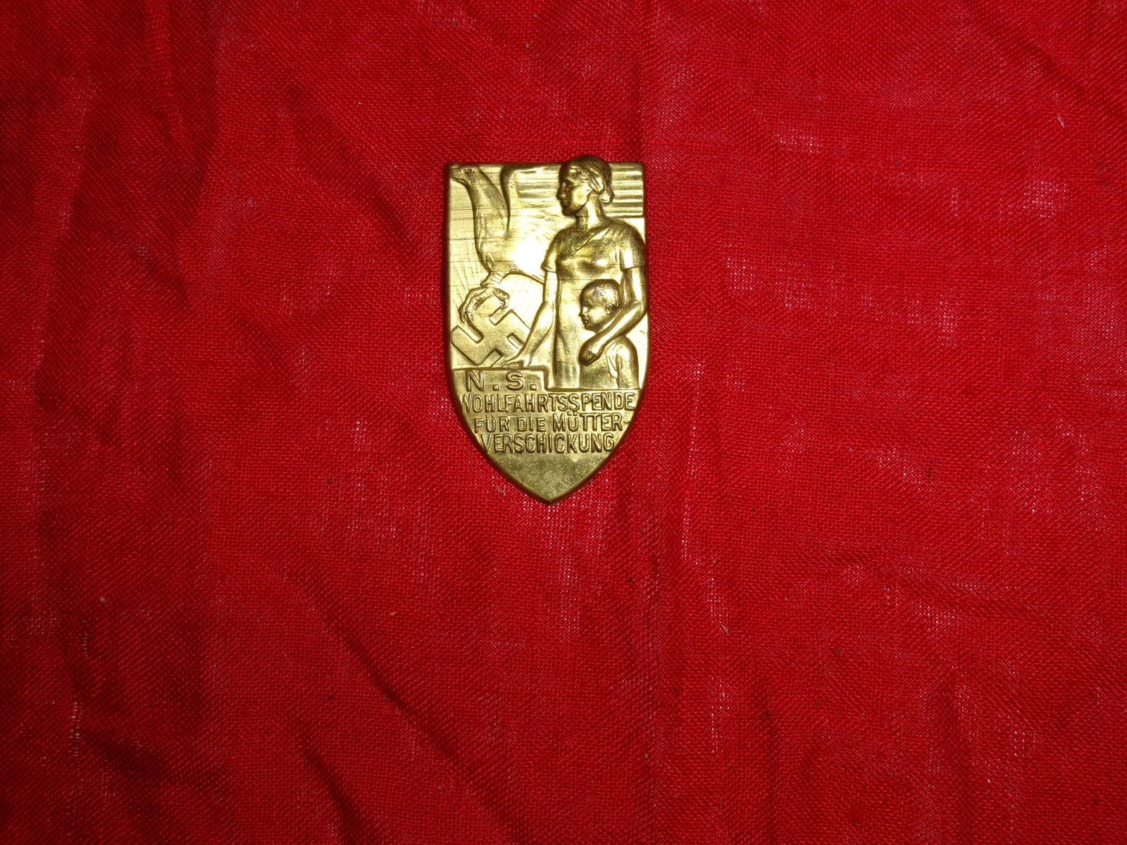 WWII German NSV Welfare Donation Badge for Mothers and Children (“Wohlfahrtsspende??): A shield-shaped gilt metal badge issued by the National Socialist People’s Welfare organization (NSV), depicting a standing mother holding a child, with a swastika and flag motif in the background. 