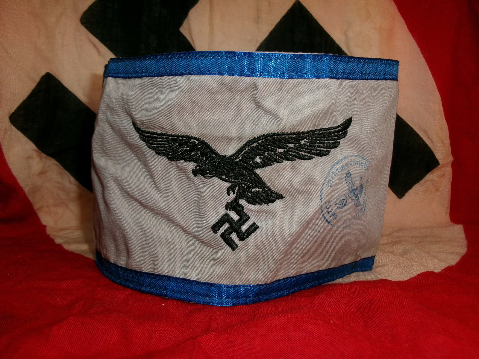 WWII German Luftwaffe Auxiliary Armband (White Variant with Blue Trim): A white cloth armband edged with blue piping, featuring an embroidered Luftwaffe eagle clutching a swastika. A faint circular ink stamp is visible on the front, likely indicating inspection or unit di