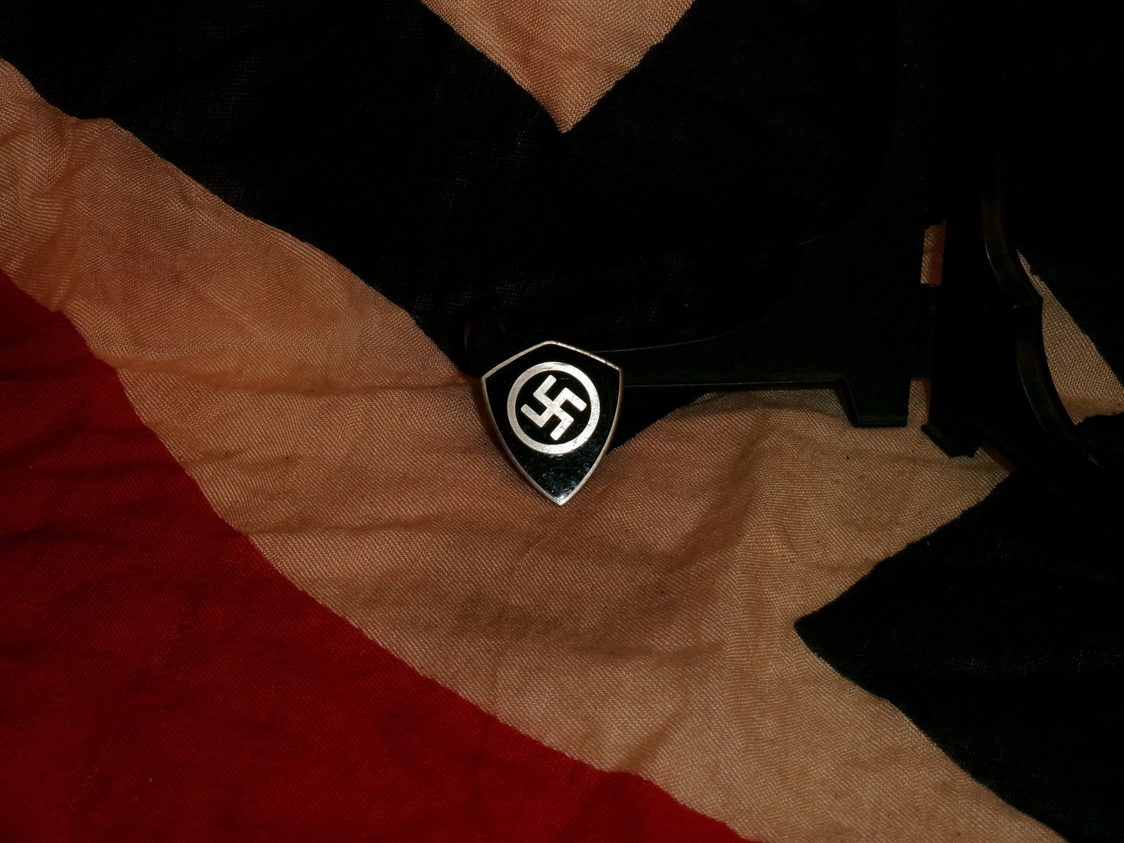 WWII German Enamel Swastika Shield Lapel Badge (“Ges. Gesch.?? Marked): A small shield-shaped metal badge with a black enamel field, centered with a white roundel and black swastika. The reverse is stamped “GES. GESCH.” (gesetzlich geschützt), indicating legal pr