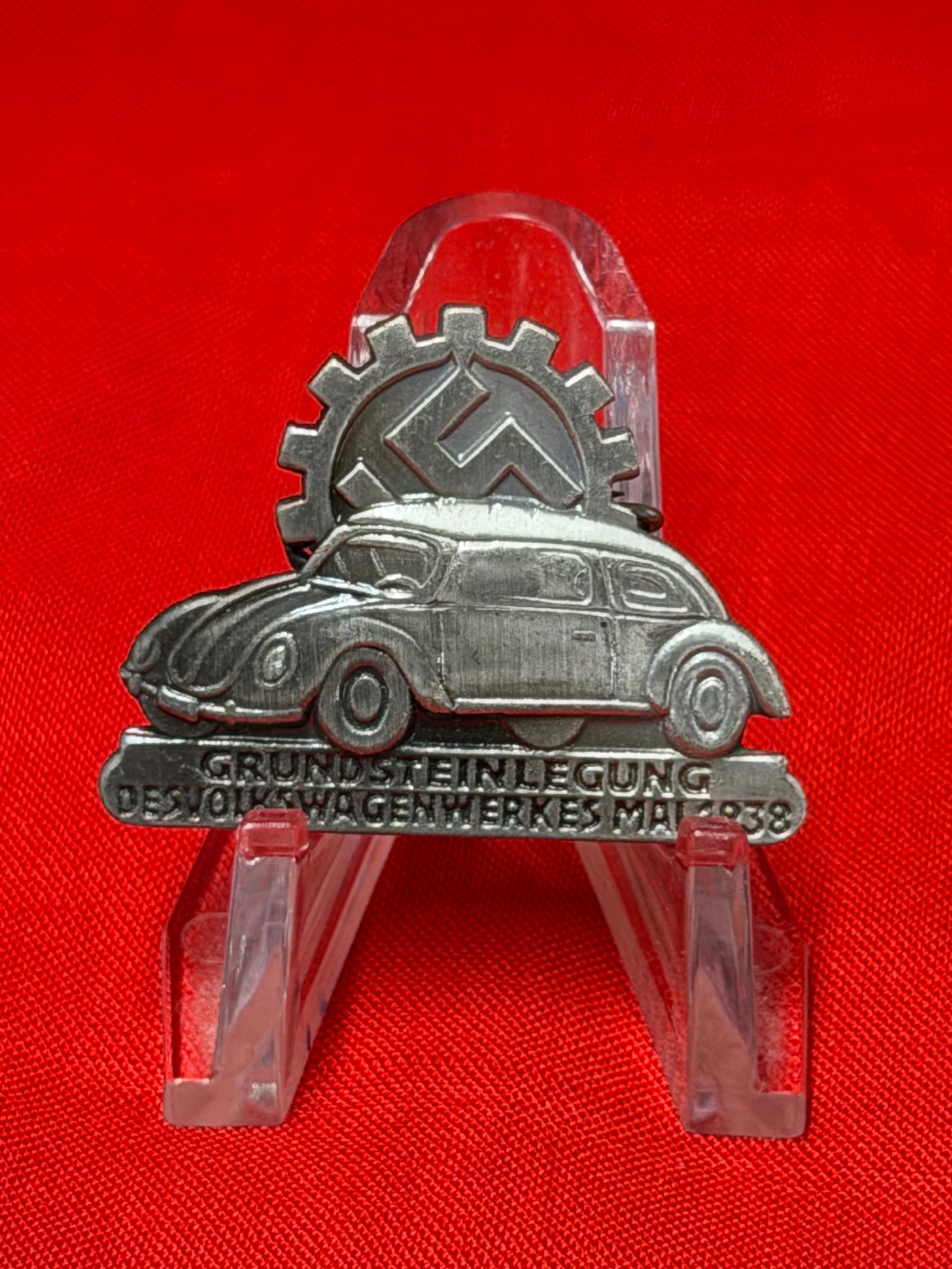 1938 German Volkswagen Factory Foundation Commemorative Pin – Fallersleben Wolfsburg: 1938 German commemorative pin featuring an early Volkswagen automobile design set before a geared emblem with stylized insignia. The lower banner reads “Grundsteinlegung des Volkswagenwerkes Mai 193