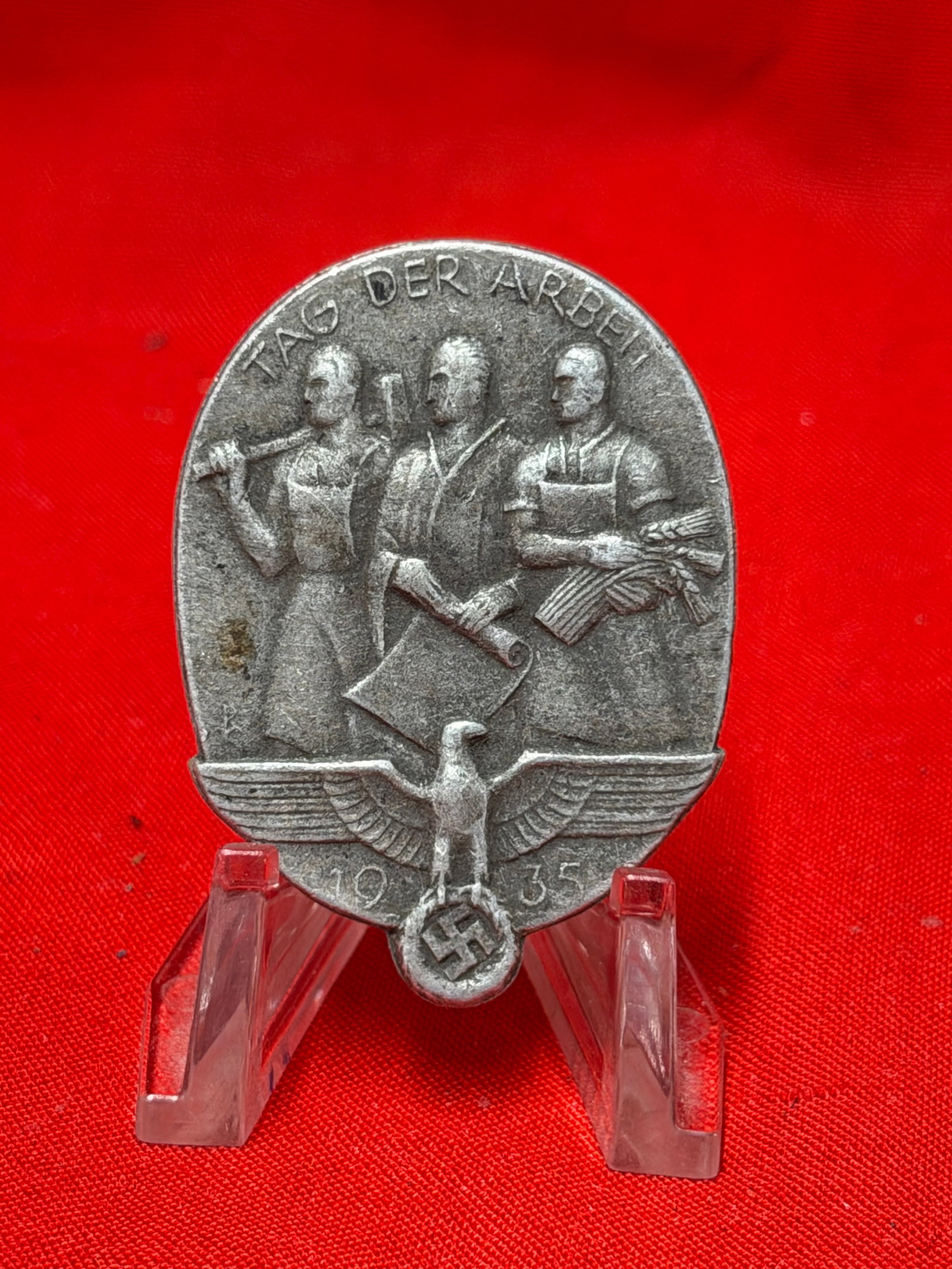 1935 German Labor Day “Tag Der Arbeit?? Metal Pin Badge – Saarlouis Marked: 1935 “Tag Der Arbeit” (Day of Labor) metal pin badge featuring detailed raised artwork with three worker figures and an eagle motif design. Reverse is marked “REDO SAARLOUIS” with an a