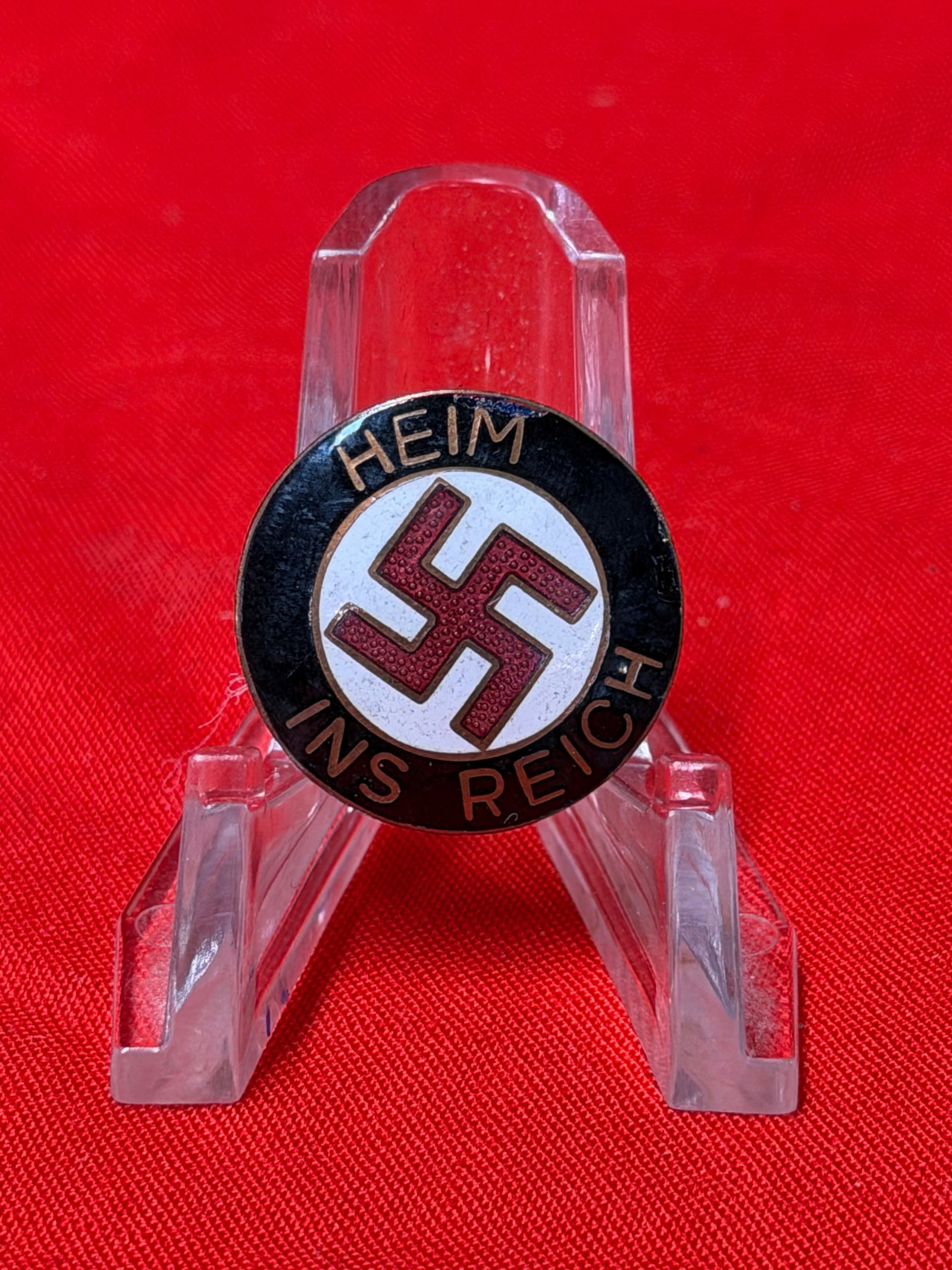 German “Heim Ins Reich?? Pin Badge WWII Era Style Political Eanameled Badge Memorabilia: Vintage round pin badge featuring the phrase “Heim Ins Reich” surrounding a central red swastika symbol on a white enamel-style background. The badge displays classic period-style German polit