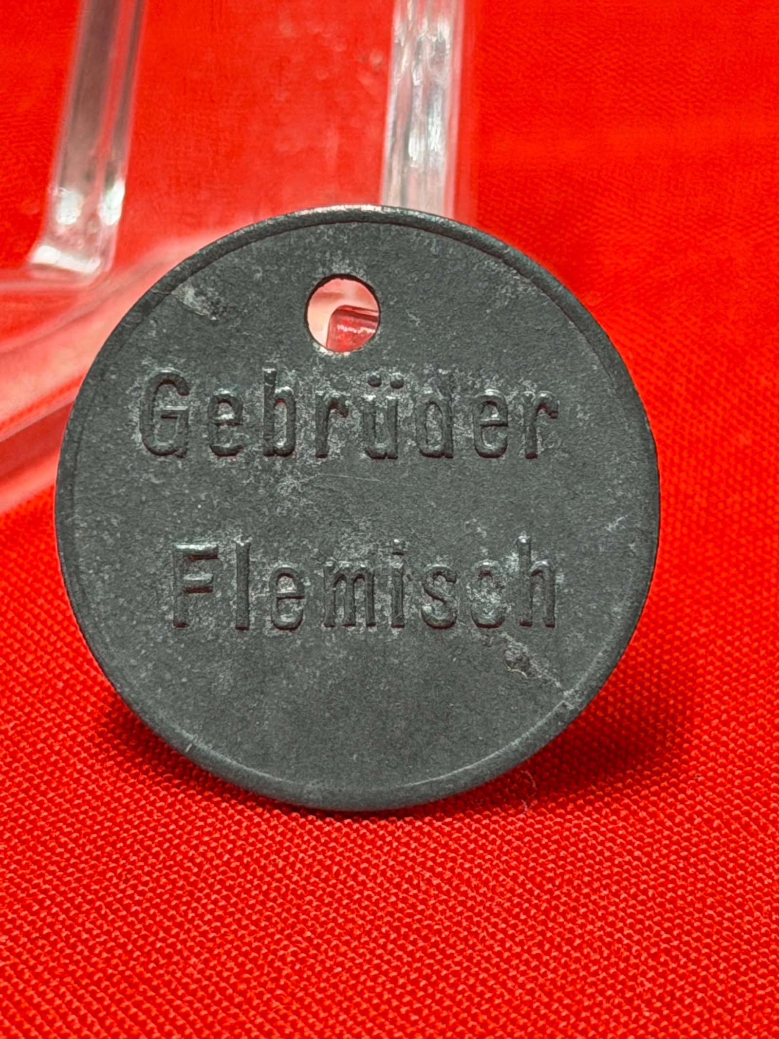 WW2 German Gebrüder Flemisch Kastenpfand Token Tag Marked 2-: This round black token tag is marked “Gebrüder Flemisch” on one side and “Kastenpfand 2-” on the reverse with a single attachment hole at the top. In our opinion, this piece appears r