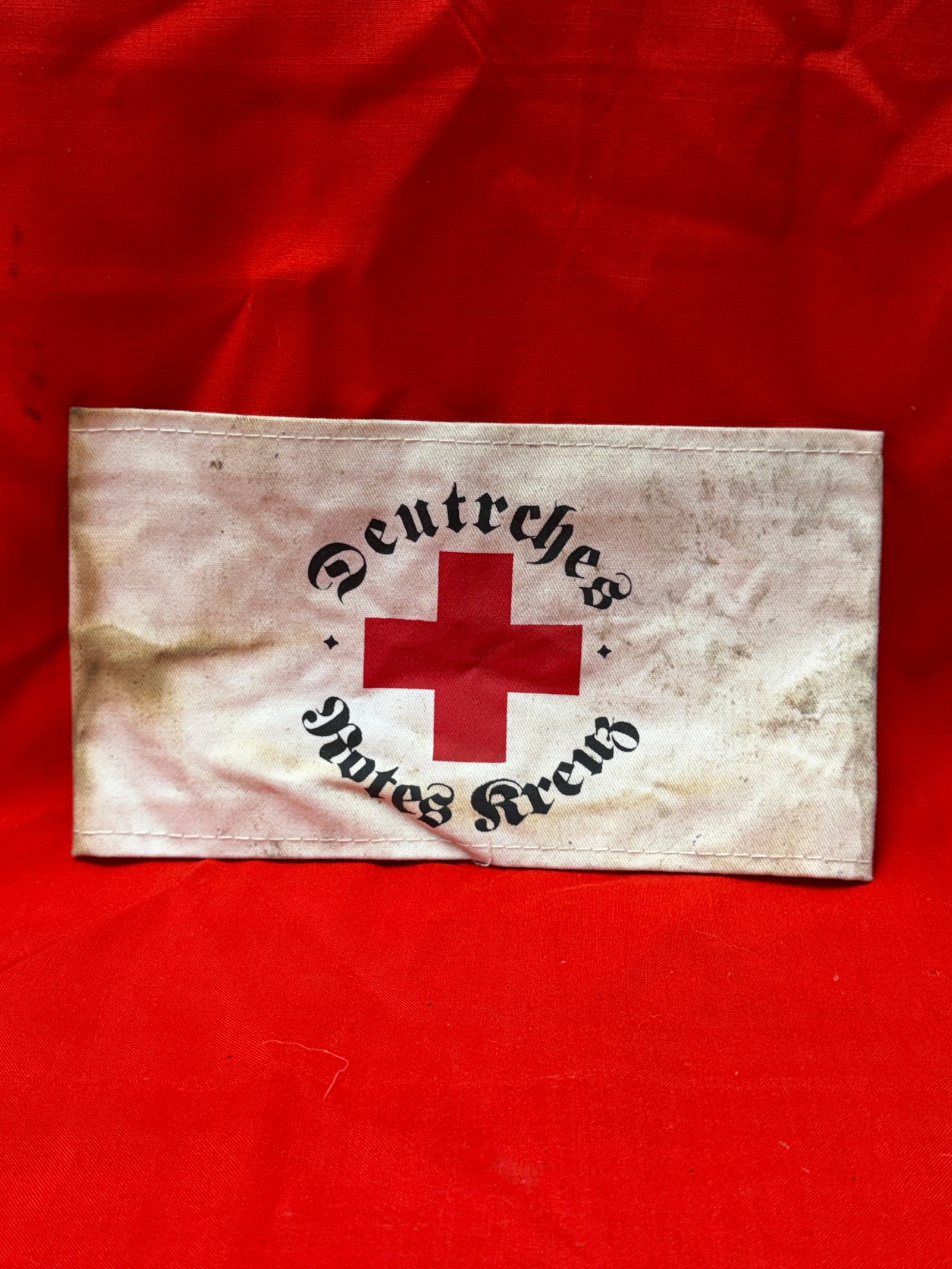 German WWII Style Deutsches Rotes Kreuz Armband Red Cross Armband: White cloth armband featuring red cross emblem with “Deutsches Rotes Kreuz” printed in black Gothic lettering across the front. Constructed from lightweight fabric with stitched edges and open
