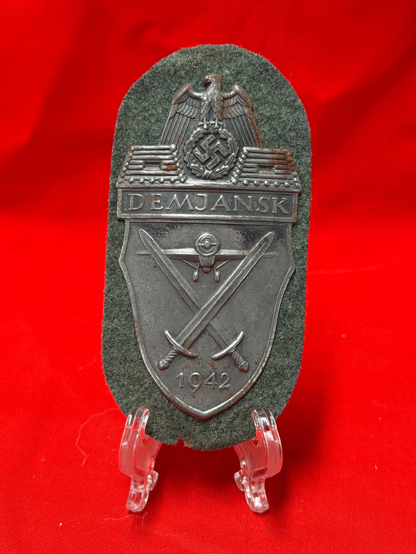 German WWII Style Demjansk Shield Patch with Metal Badge on Wool Backing: Metal Demjansk campaign-style shield mounted on green wool backing featuring eagle, crossed swords, aircraft motif, and 1942 date detail. Reverse shows paper backing with age-related wear, creasing, a