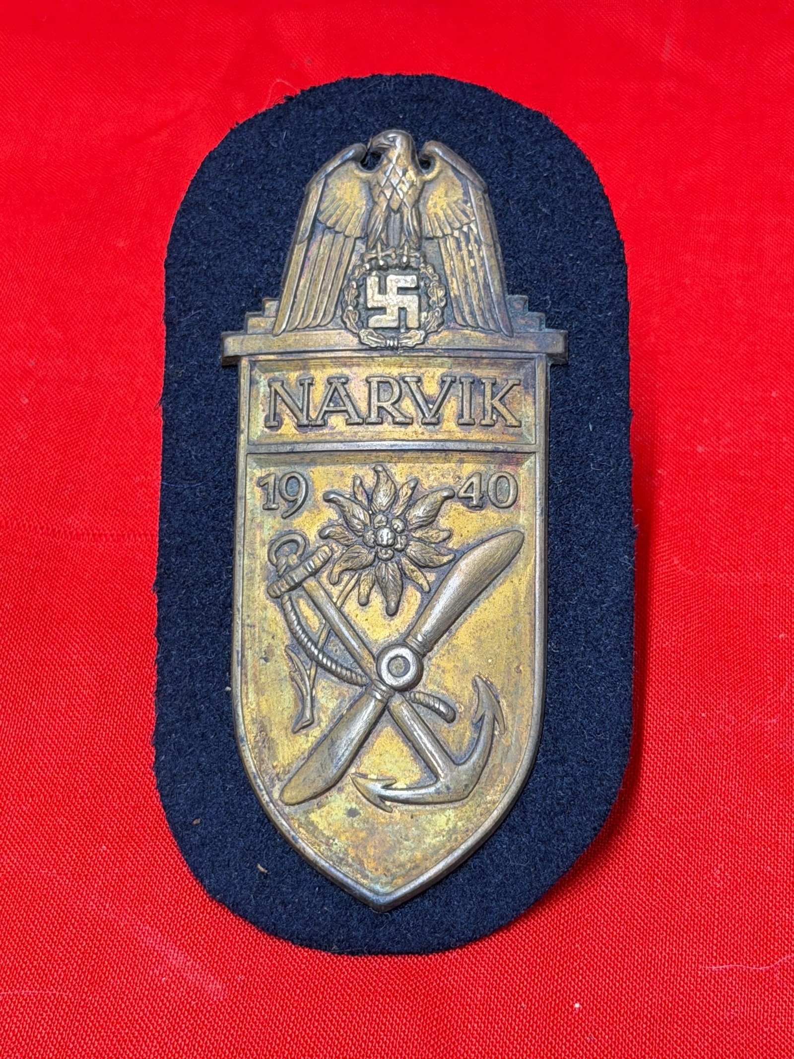 WW2 German Narvik 1940 Campaign Shield Cloth Backed Badge: Narvik 1940 campaign shield badge mounted on dark cloth backing featuring eagle, anchor, propeller, and edelweiss design elements. In our opinion, this piece appears to be a German military commemorat