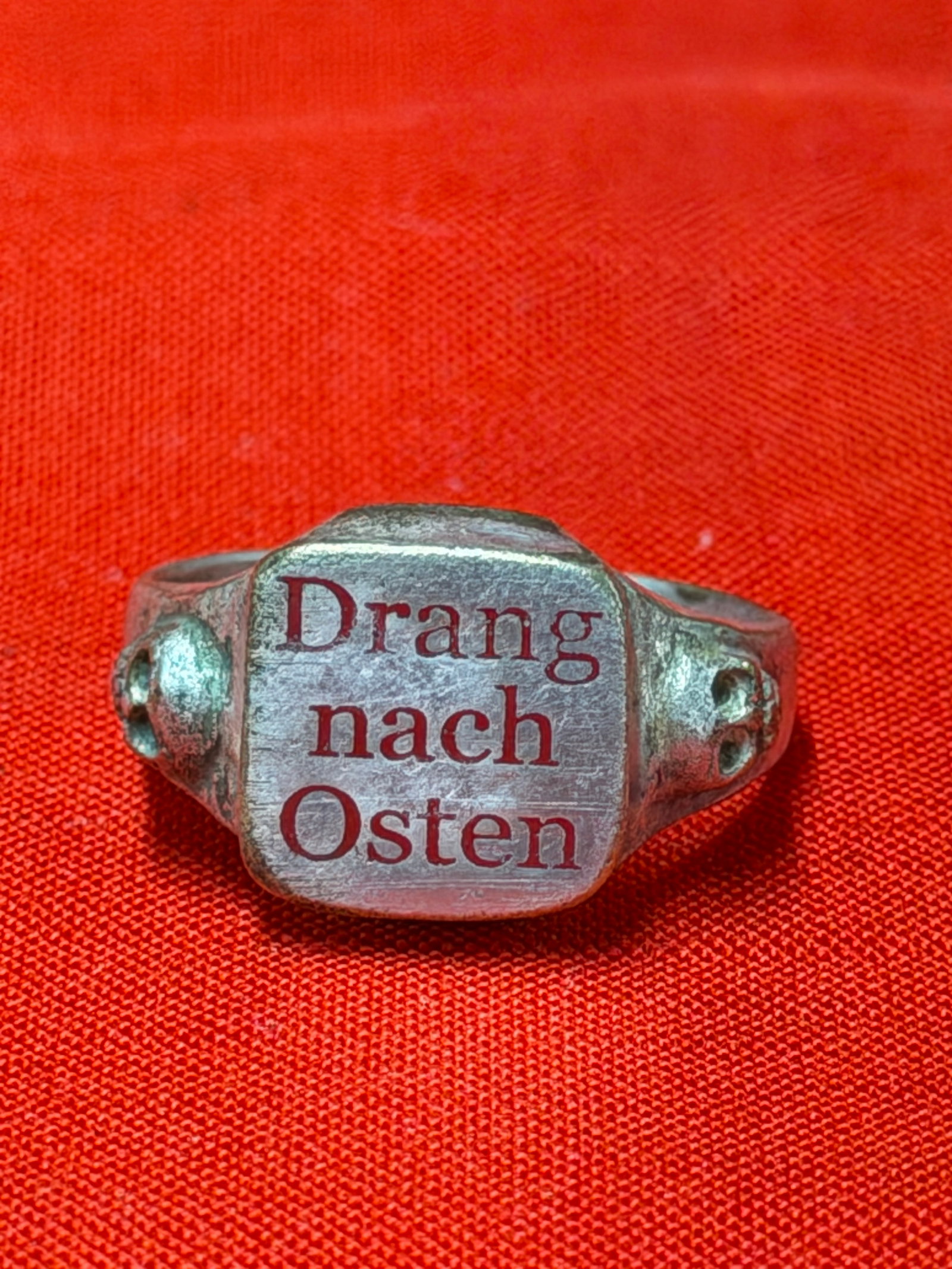 WW2 German “Drang Nach Osten?? Totenkopf SS Skulls Silver Tone Ring: WW2 German “Drang Nach Osten” style silver-tone ring featuring engraved red lettering on the face and skull-style side details. Ring displays visible age, patina, finish wear, and surface mark
