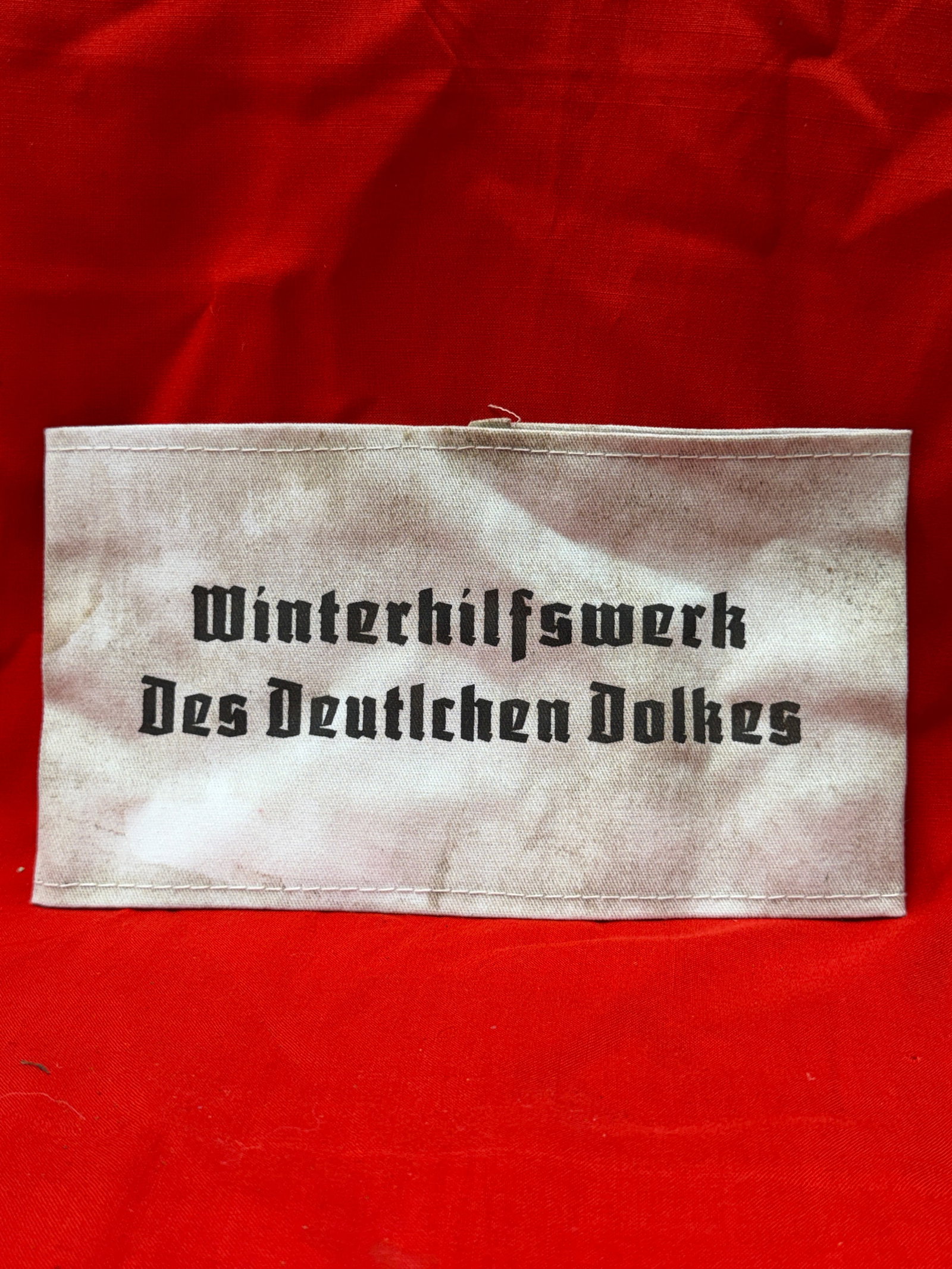 WW2 German Winterhilfswerk Des Deutschen Volkes Cloth Armband Sleeve: Winterhilfswerk Des Deutschen Volkes cloth armband sleeve featuring black German script lettering on a light fabric background. Piece shows age-related wear including staining, discoloration, loose th
