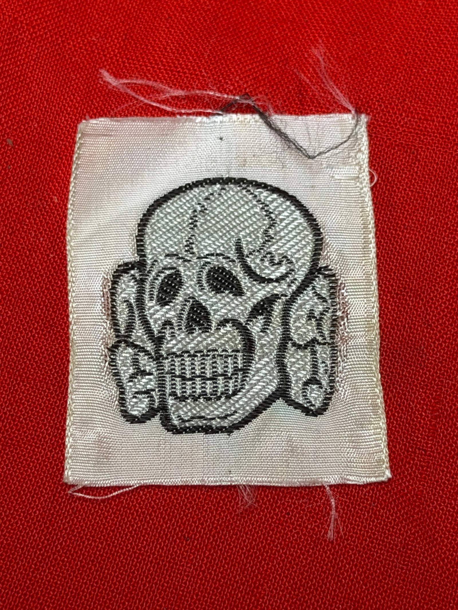 WWII German Totenkopf Skull Cloth Insignia Patch: WWII era German Totenkopf (death’s head skull) style cloth insignia patch featuring embroidered skull design on light colored fabric backing. Small size example likely intended for uniform or cap ap