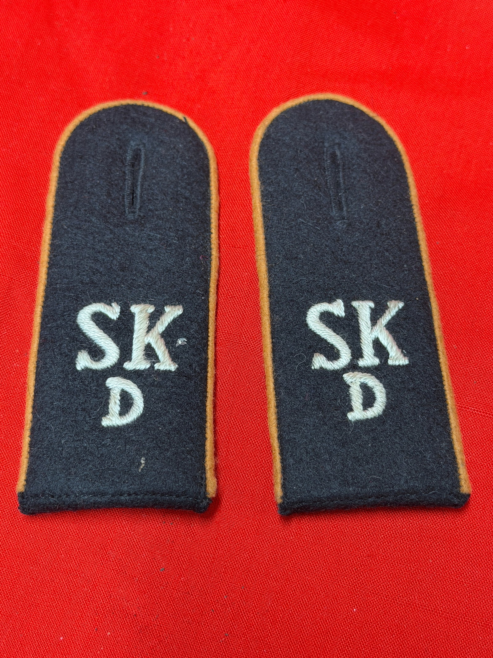 Pair of WWII German SKD Uniform Shoulder Boards / Slip-On Epaulettes: WWII German uniform shoulder boards / slip-on epaulettes featuring embroidered “SKD” lettering on dark wool backing with tan/gold piping around the edges. These appear to be period military or