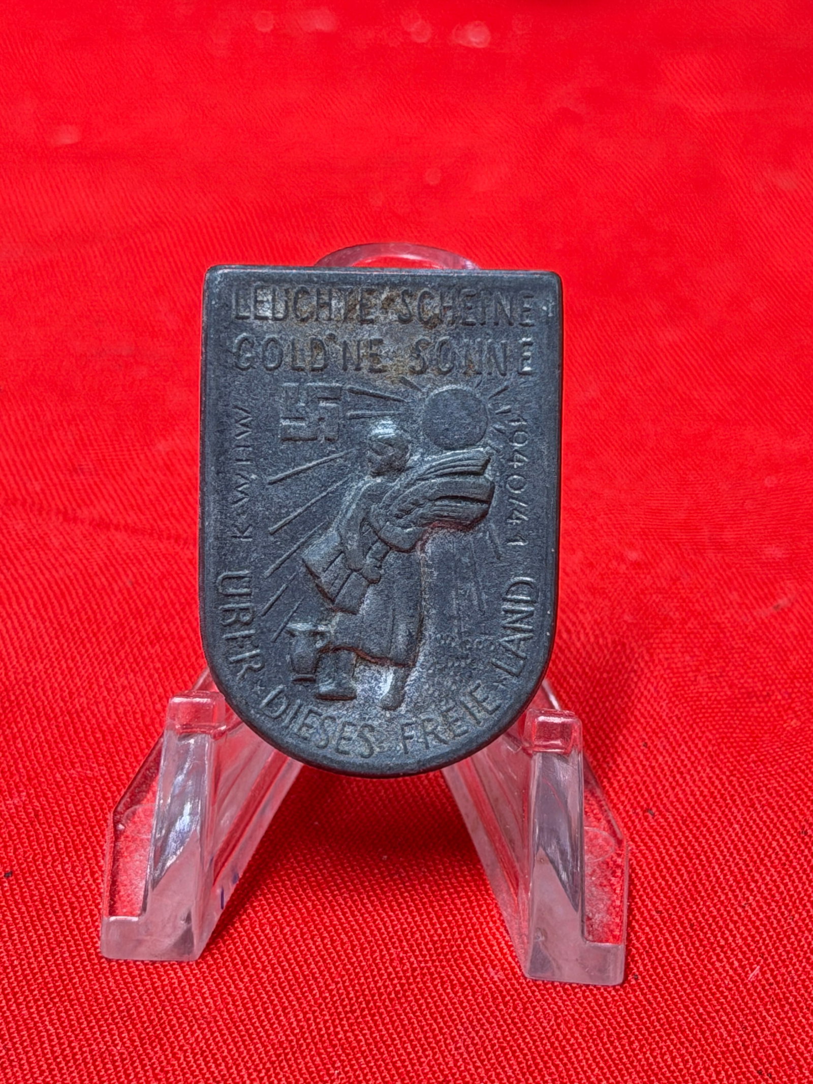 WWII German Tinnie Badge – “Leuchtende Gold’ne Sonne?? Propaganda/Event Pin: WWII-era German tinnie (membership/event badge) featuring the slogan “Leuchtende Gold’ne Sonne” with a stylized sunburst and figure motif on the front. The badge includes additional German t