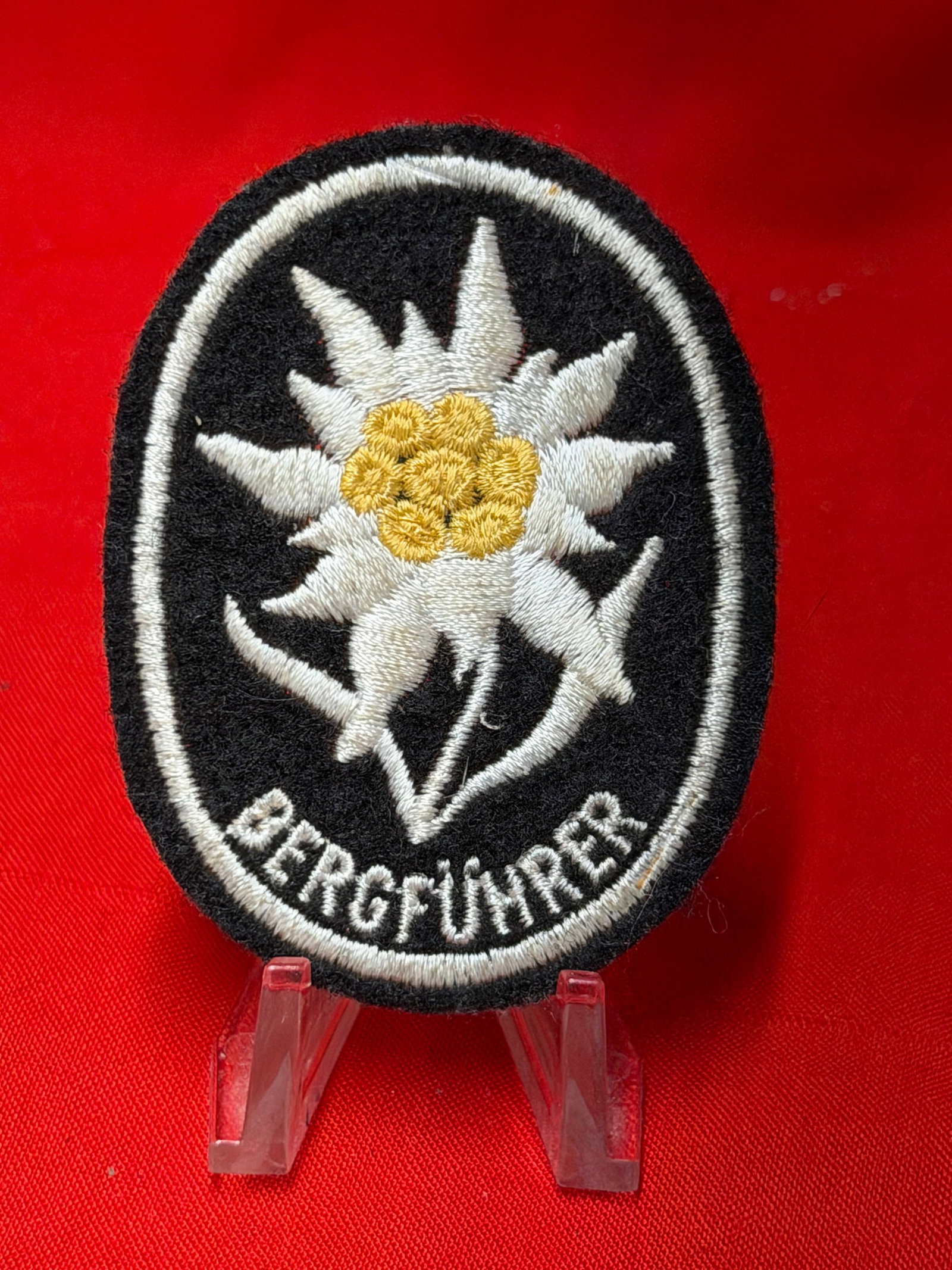 WWII German Bergführer Edelweiss Mountain Guide Cloth Sleeve Patch: WWII German Bergführer cloth insignia patch featuring embroidered Edelweiss flower design on black wool backing with “BERGFÜHRER” lettering along the lower edge. White and gold thread embroi