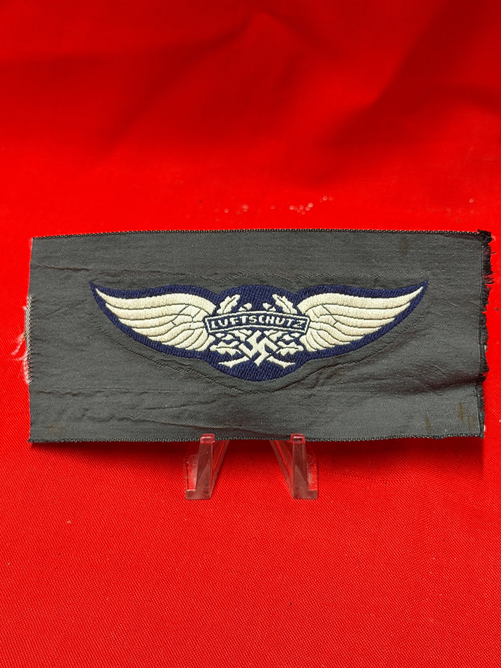 WWII German Luftschutz Embroidered Cloth Wing Insignia Patch: German Luftschutz embroidered cloth insignia featuring winged emblem with “LUFTSCHUTZ” banner across the center. Blue and white stitched design mounted on dark gray cloth backing. Measures app