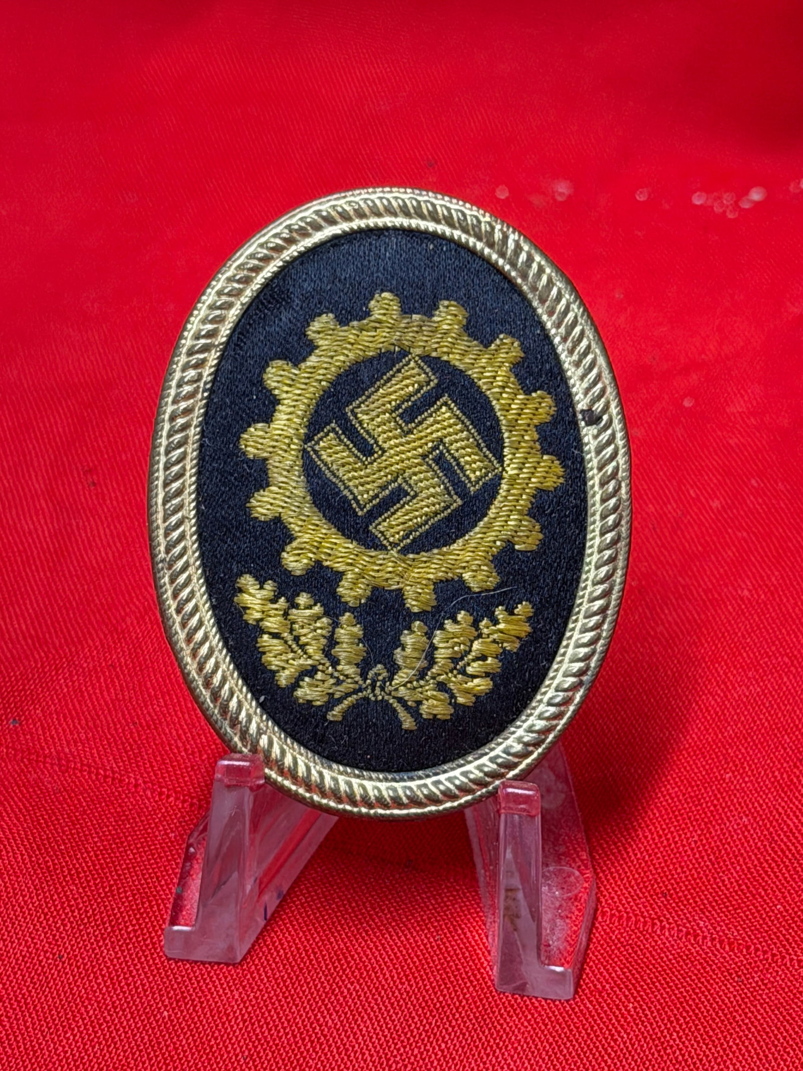 WWII German RAD Reichsarbeitsdienst Oval Uniform Tinnie Badge M1/357: German RAD related oval uniform badge featuring embroidered gearwheel emblem with central swastika and oakleaf detail on dark cloth backing with metal frame. Reverse marked “Lieferant No 357” 