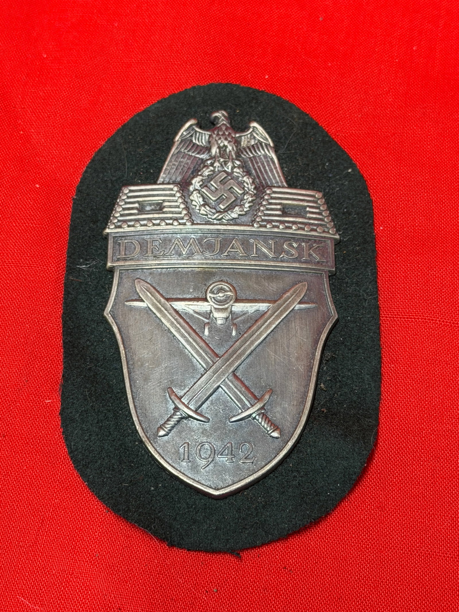 WWII German Demjansk Campaign Shield 1942 Badge on Black Cloth Backing: German Demjansk campaign shield badge marked “DEMJANSK 1942” featuring eagle crest, crossed swords, and aircraft motif mounted on black cloth backing. In our opinion, this piece follows the Wo