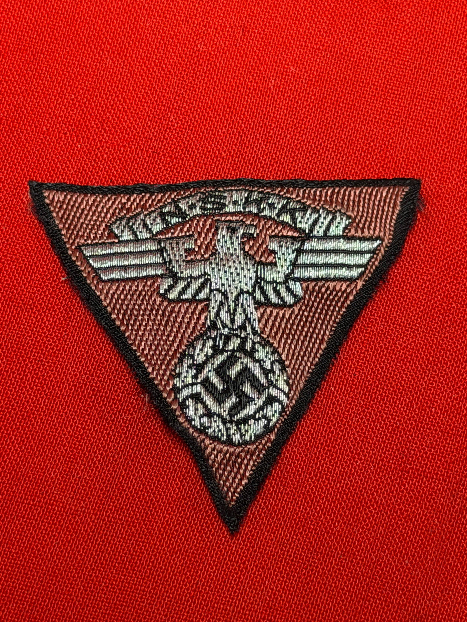 WWII German NSKK Cap Eagle Insignia Patch – National Socialist Motor Corps: Second World War German NSKK (Nationalsozialistisches Kraftfahrkorps) cap insignia patch featuring silver-gray embroidered national eagle with swastika on brown triangular backing with black edge bord