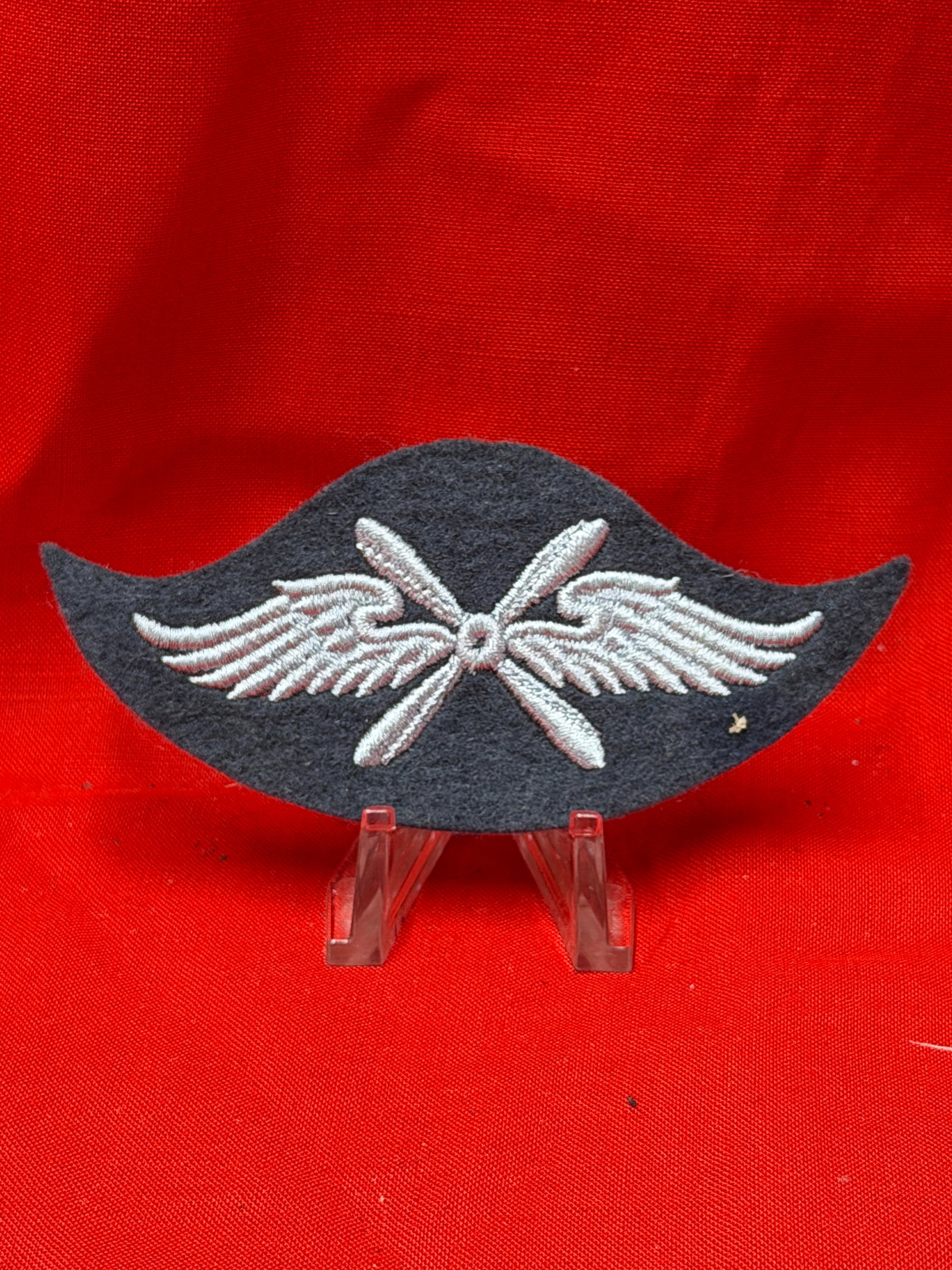 WWII German Luftwaffe Propeller and Wings Cloth Insignia Patch: This German Luftwaffe themed cloth insignia patch features embroidered silver colored wings with crossed propeller blades on a dark wool backing. The unique curved shape and detailed stitching give th