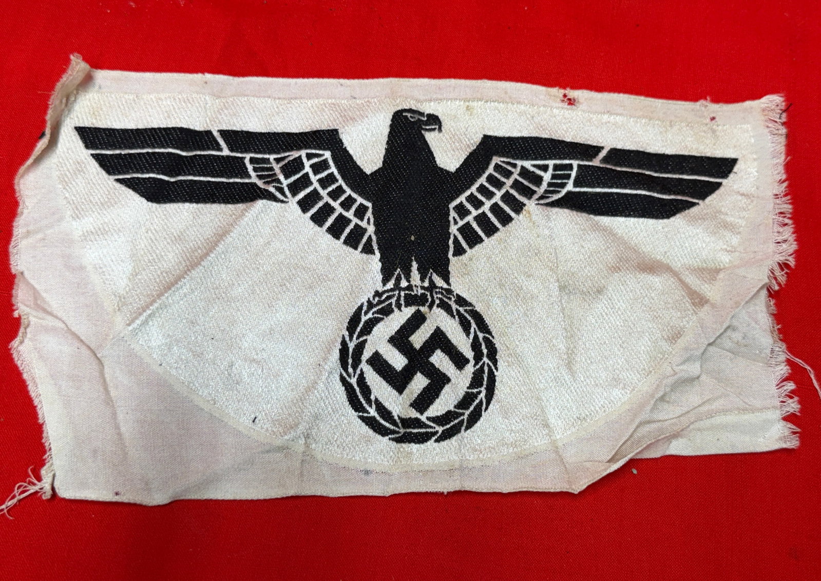 German Eagle BeVo Type Cloth Insignia Panel WWII Era Memorabilia: This German eagle themed cloth insignia panel features a black and white woven design with eagle and wreath imagery on a light fabric backing. The piece displays detailed woven construction with unfin