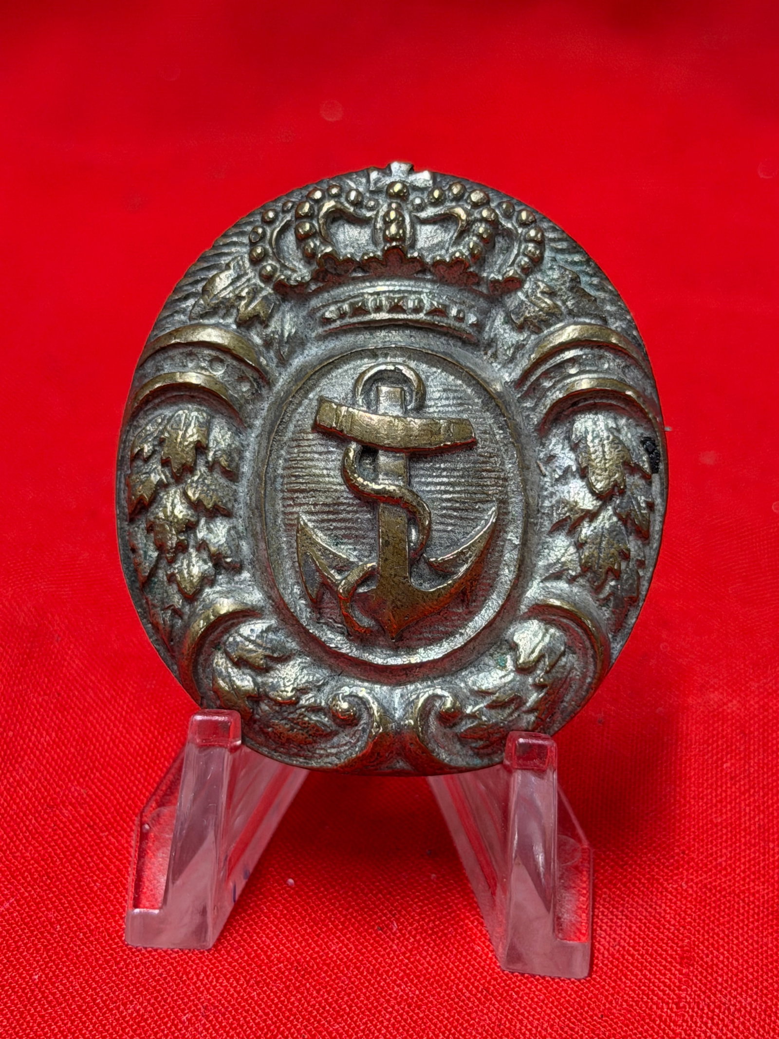 Imperial German Naval Anchor Crown Metal Badge Pin: This Imperial German naval themed metal badge features a crowned oval design with a central anchor emblem and decorative leaf detailing around the border. The piece displays a two tone appearance with