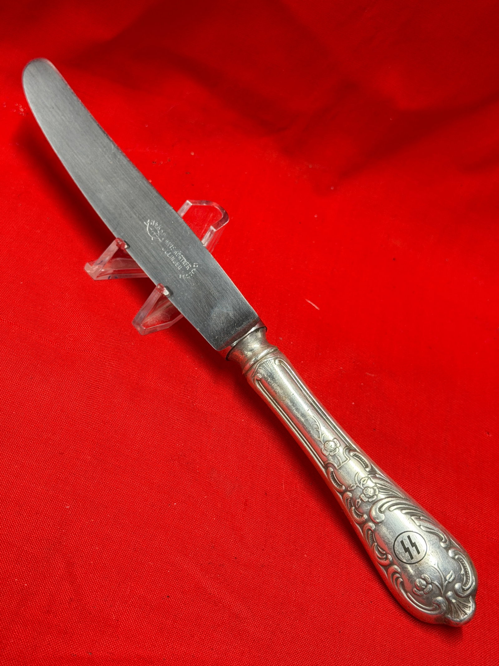 WWII German SS Marked Table Knife – Hans Stortz Solingen Stainless Steel: WWII era German table knife featuring ornate silver-toned floral pattern handle with SS runes emblem inset at the terminal. Blade is maker marked “Hans Stortz Solingen” with stainless marking 