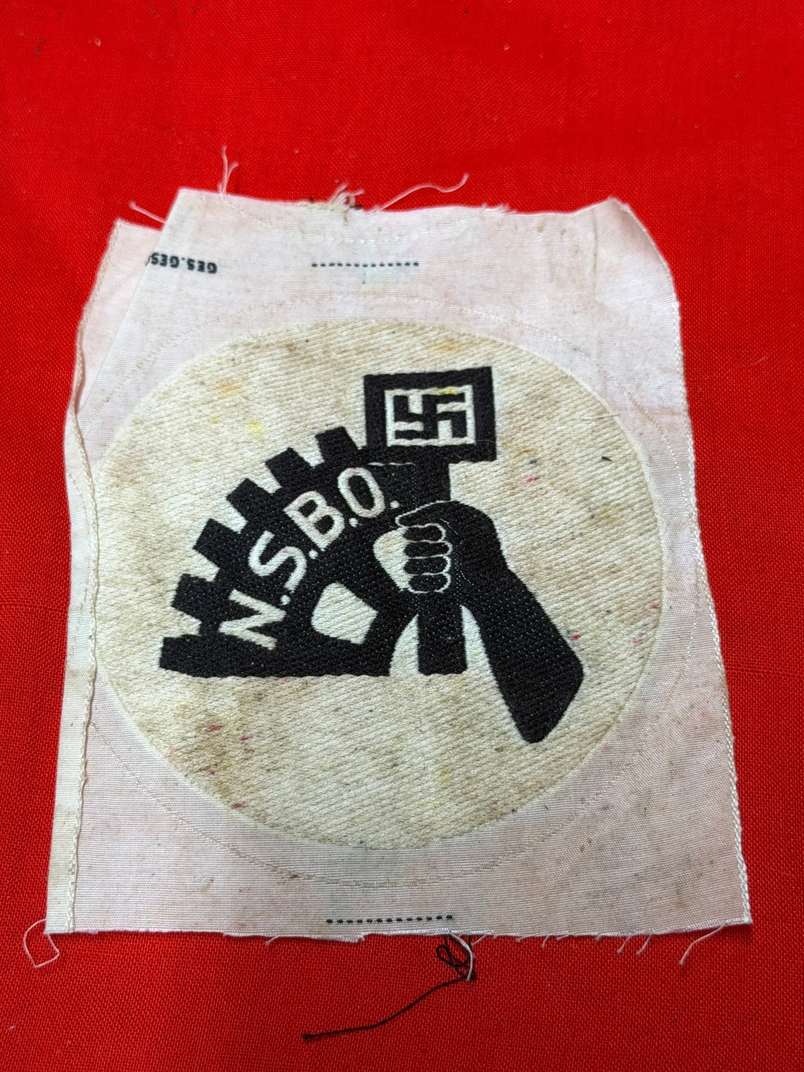WWII German NSBO Cloth Insignia Patch – National Socialist Factory Cell Organization: WWII era German NSBO (Nationalsozialistische Betriebszellenorganisation) cloth insignia patch featuring black embroidered emblem with gearwheel, flag, swastika, and “N.S.B.O.” lettering on whi