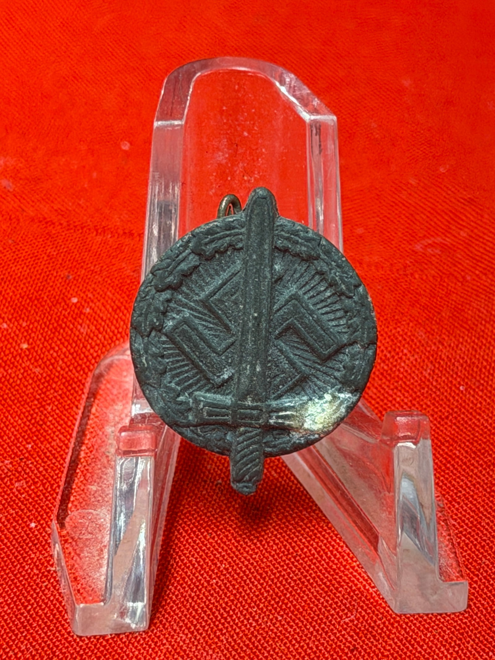 WWII German Tinnie Badge With Sword Motif: World War II era German tinnie badge featuring circular wreath design with central sword and surrounding emblem details. In our opinion, the piece is constructed from darkened lightweight metal with f