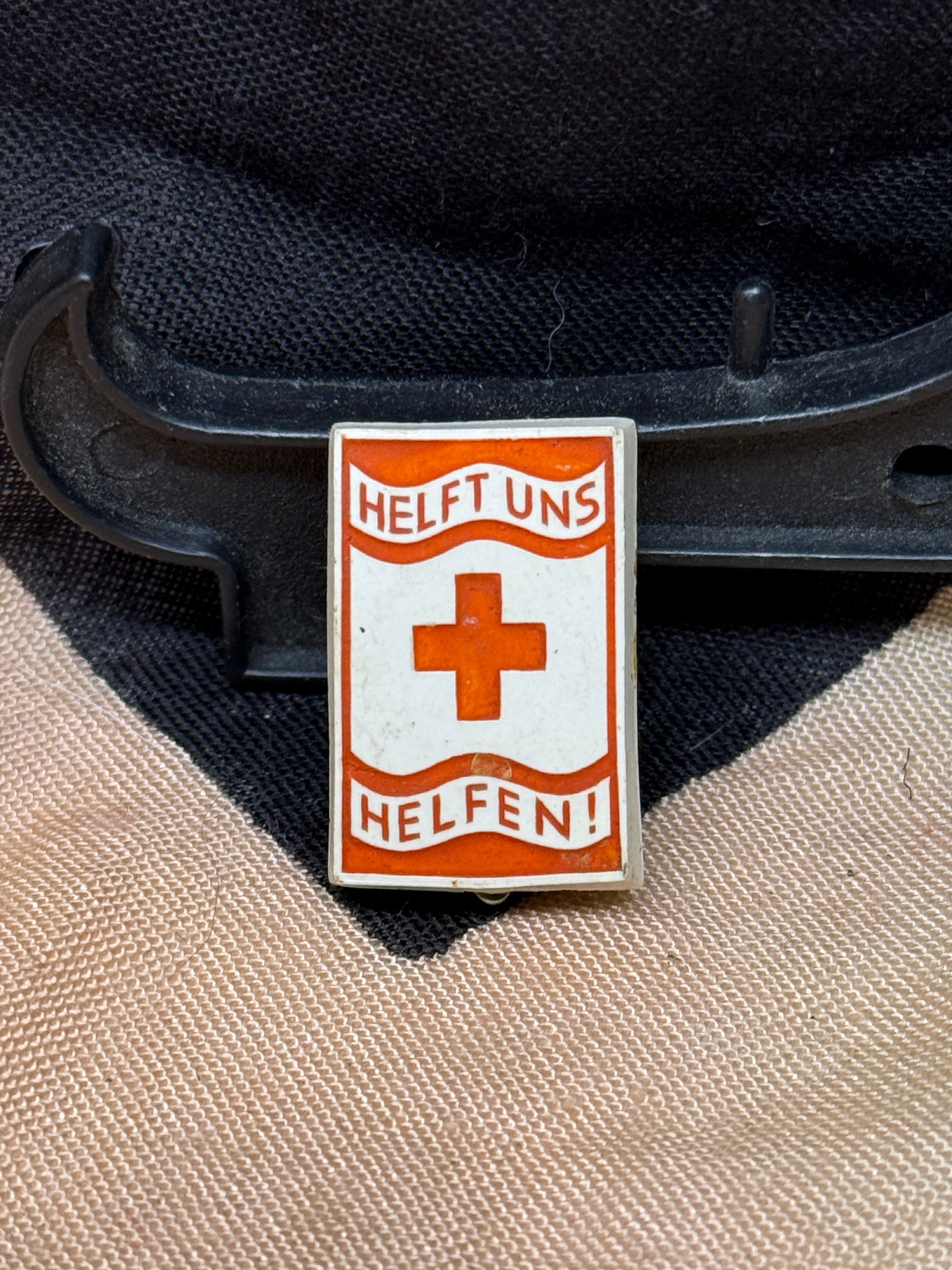 WWII German Red Cross “Helft Uns Helfen!?? Relief Support Pin: This rectangular enamel pin bears the slogan “HELFT UNS HELFEN!” (“Help Us Help!”) surrounding a red cross emblem on a white background with red borders. Produced during the World War 