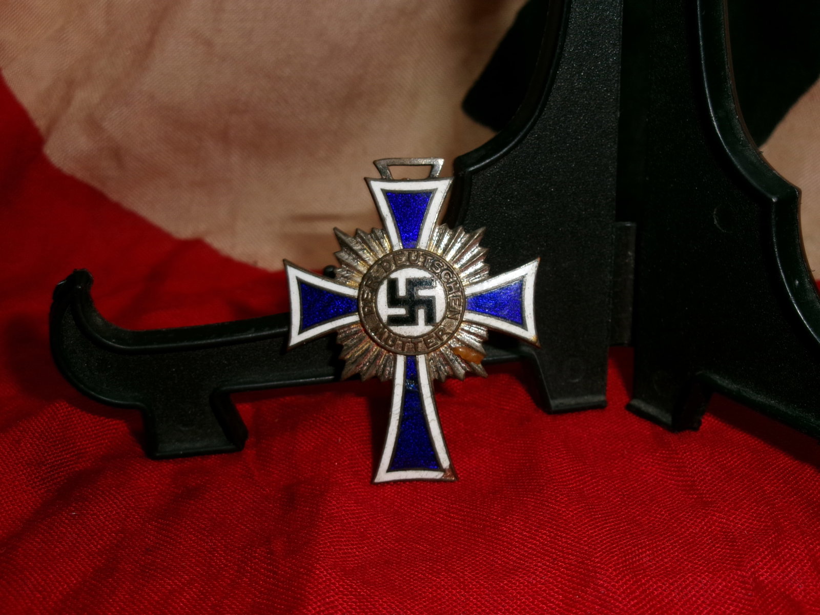 WWII German Mother’s Cross (Ehrenkreuz der Deutschen Mutter), Bronze Class: World War II German Mother’s Cross award featuring blue and white enamel cross arms with central black swastika surrounded by the inscription “DER DEUTSCHEN MUTTER.” Reverse engraved “16 D