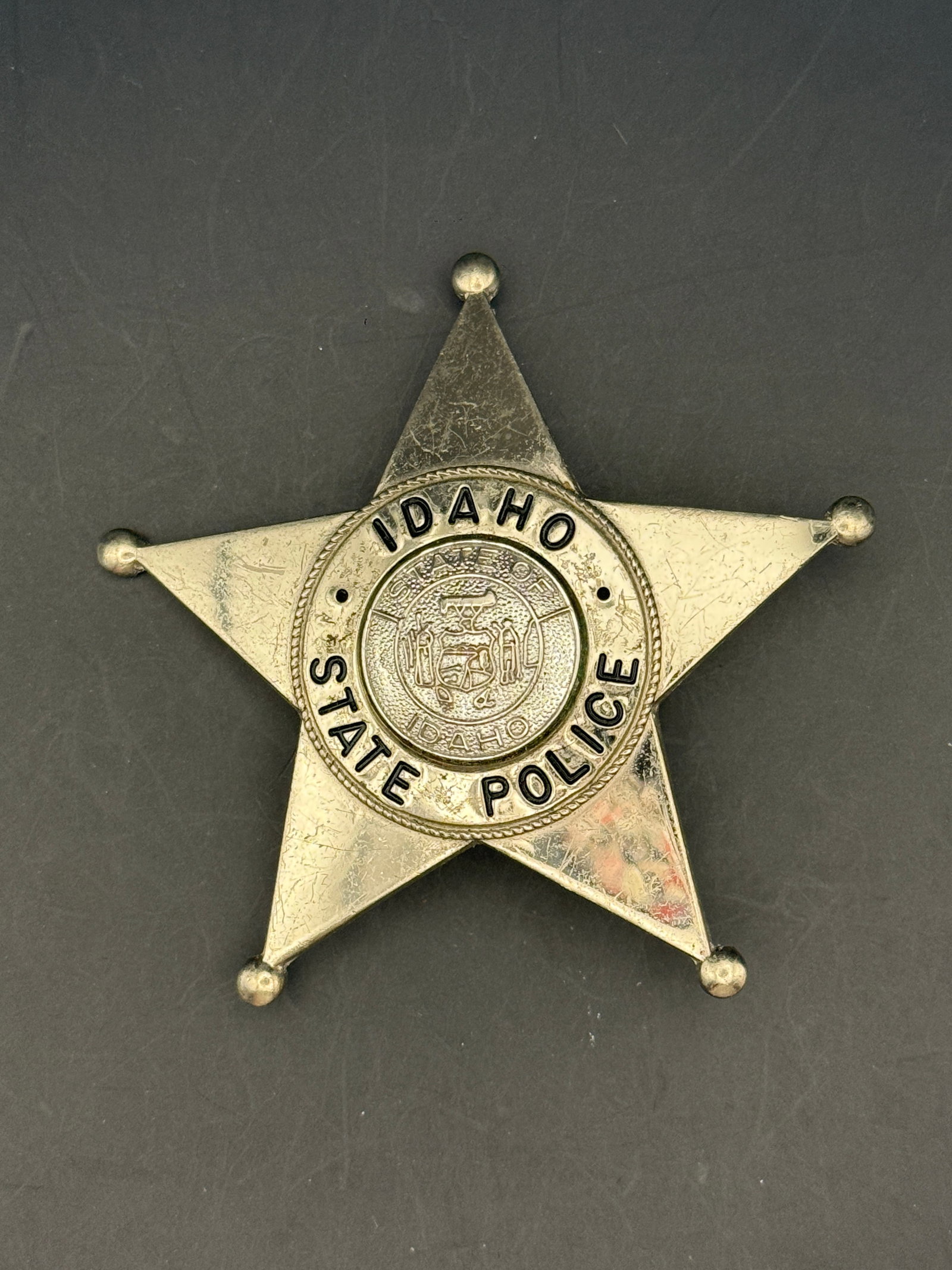 Mid-20th Century Idaho State Police Badge by Ga-Rel: Five-point star-shaped law enforcement badge bearing the inscription “IDAHO STATE POLICE” surrounding the official Idaho state seal at center. Manufactured in silver-toned metal with raised le