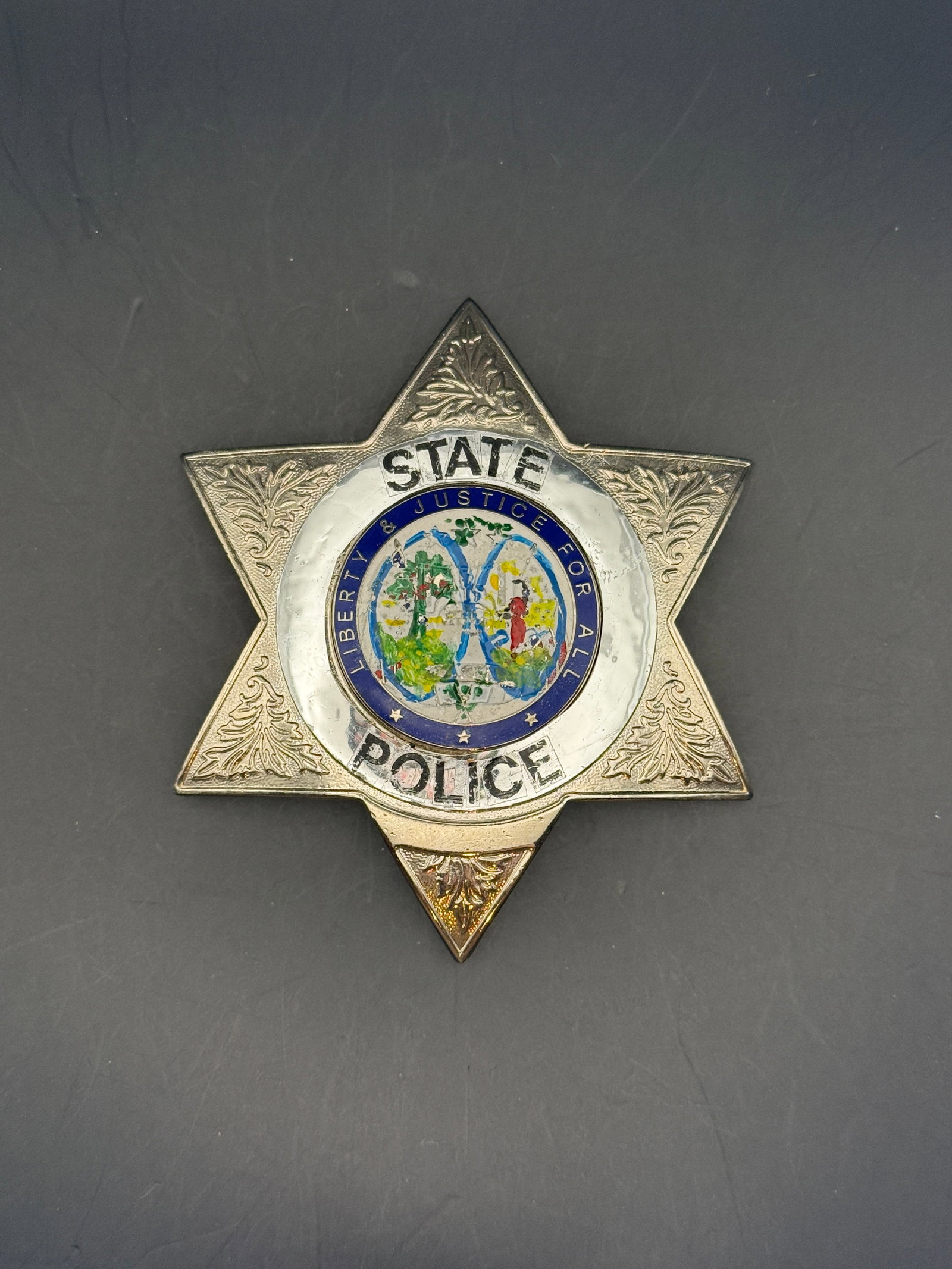 Mid-20th Century State Police Badge: Six-point star-shaped State Police badge featuring engraved decorative flourishes and a central multicolored seal bearing the motto “Liberty & Justice For All.” The badge is constructed in sil