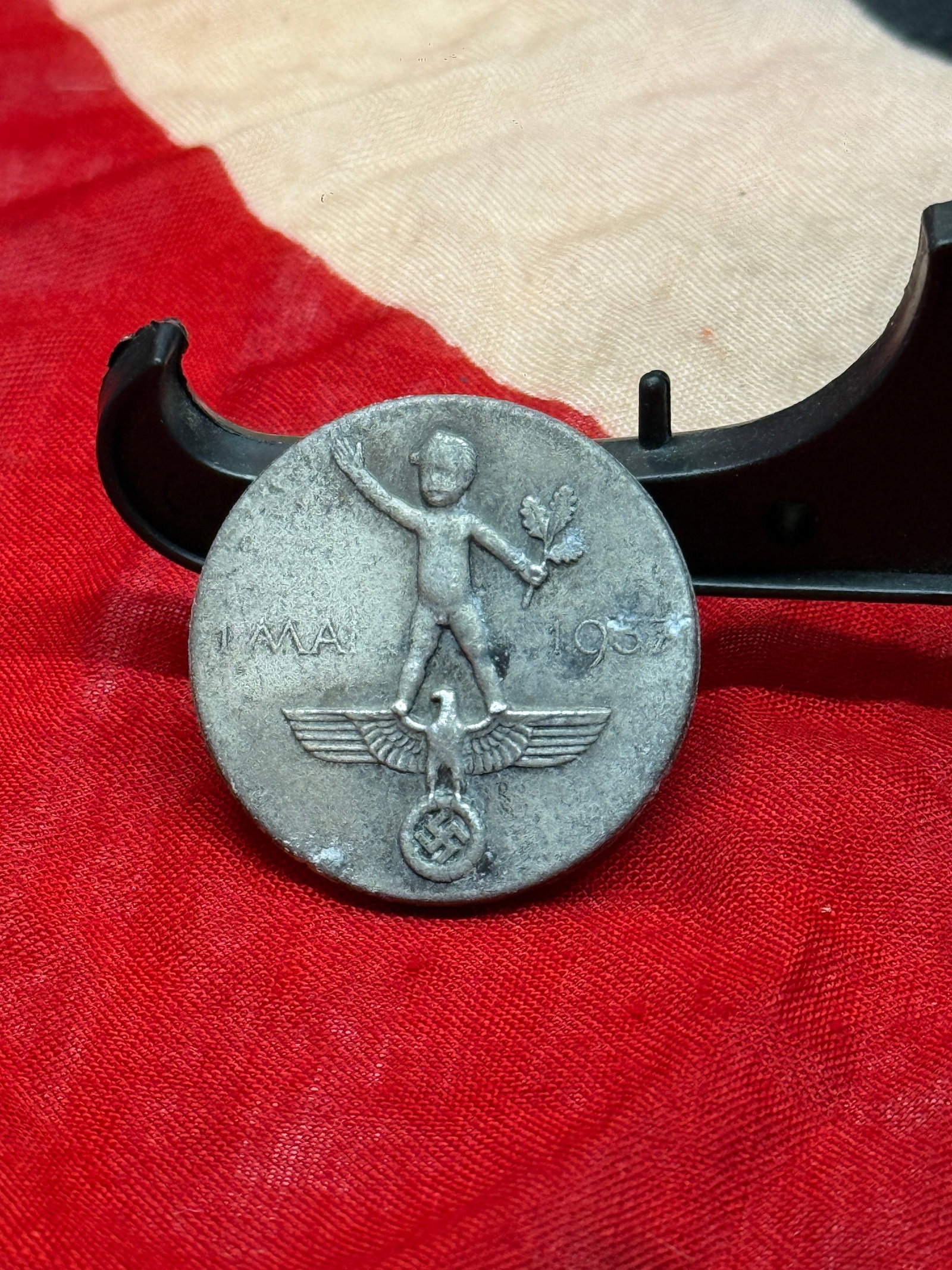 1937 German National Day of Labor Tinnie Badge: German 1937 Tag der Nationalen Arbeit (National Day of Labor) commemorative tinnie badge depicting a child figure holding an oak branch while standing atop a Reich eagle and swastika emblem. Issued in