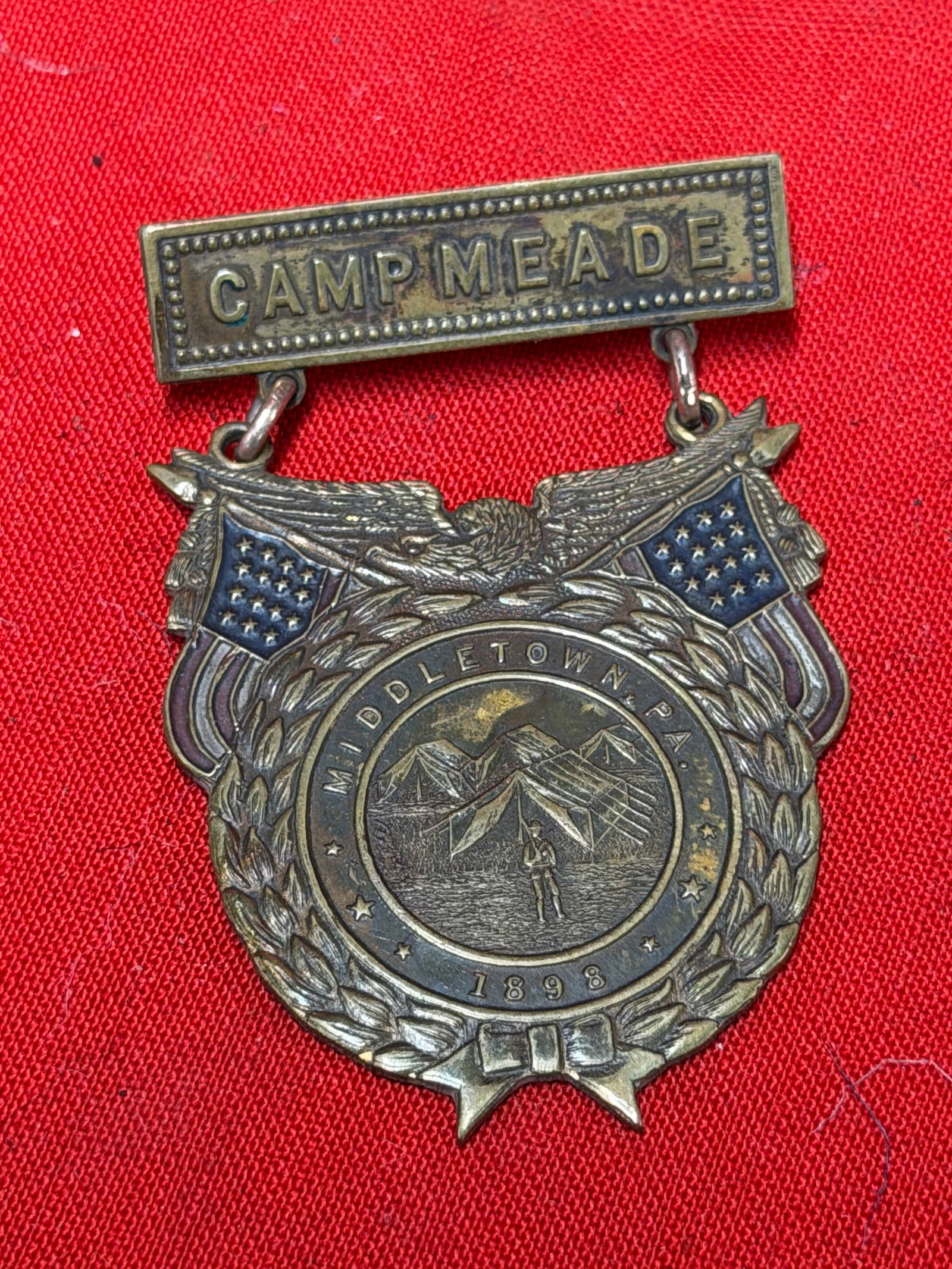 1898 Camp Meade Middletown PA Spanish American War Patriotic Badge Pin C.M. Robbins: Offered for sale is a decorative commemorative badge featuring a top bar marked “Camp Meade” and a suspended shield-shaped medal with patriotic motifs including crossed American flags, an eagl