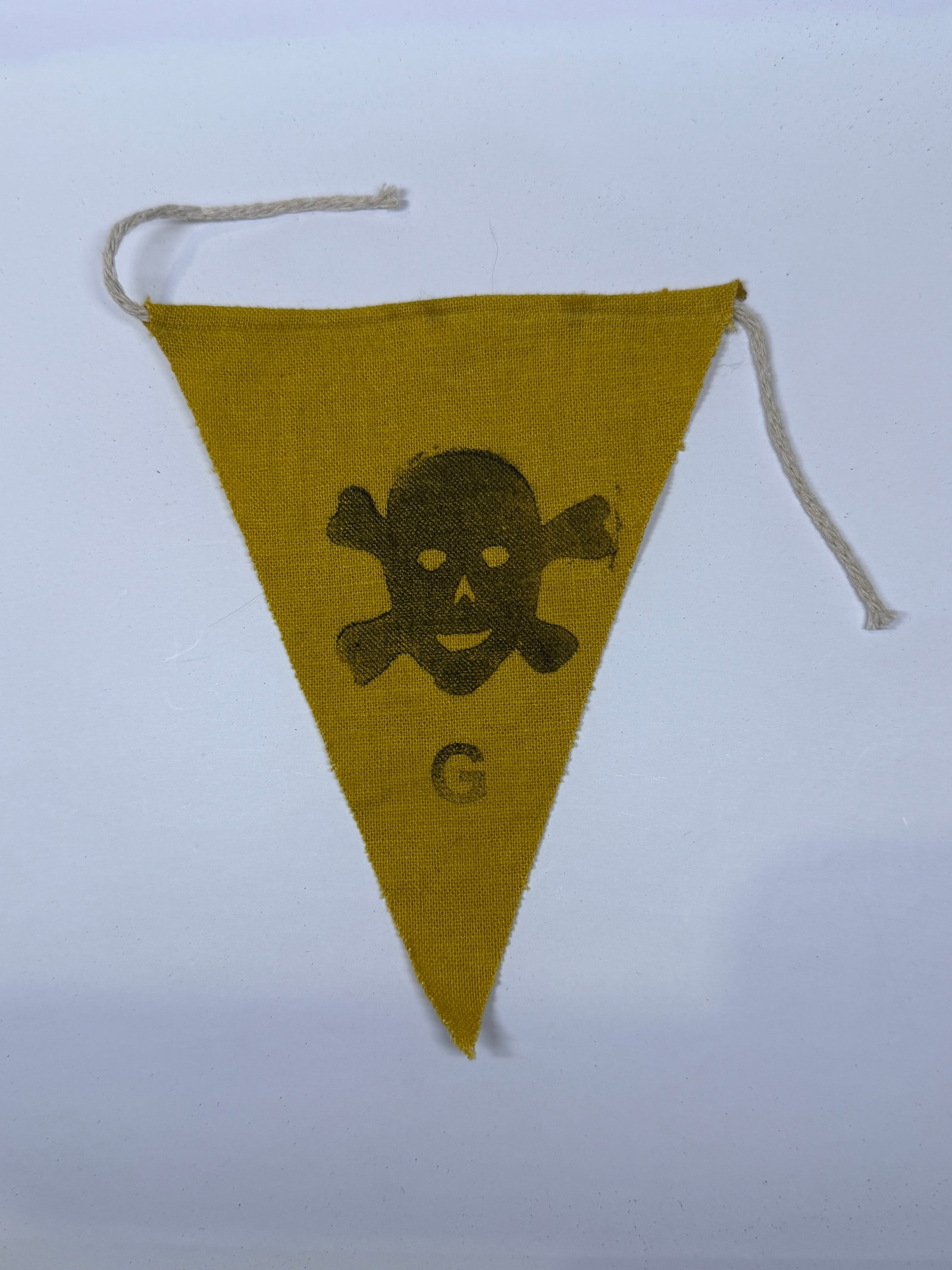 WWII German Skull-and-Crossbones Warning Pennant: This small triangular yellow cloth pennant features a prominently printed skull-and-crossbones motif with a letter “G” beneath the design. Equipped with attachment cords, the pennant was inten
