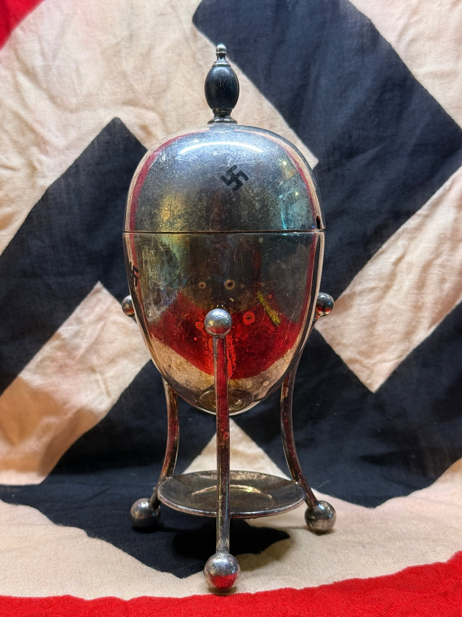 WWII Era German Silver-Plated Covered Condiment Pot with Swastika Motif and Stand: Silver-plated covered condiment pot featuring a domed lid with finial, decorative four-foot stand, and swastika motifs applied to the exterior of the lid. The vessel includes a spoon notch at the rim,
