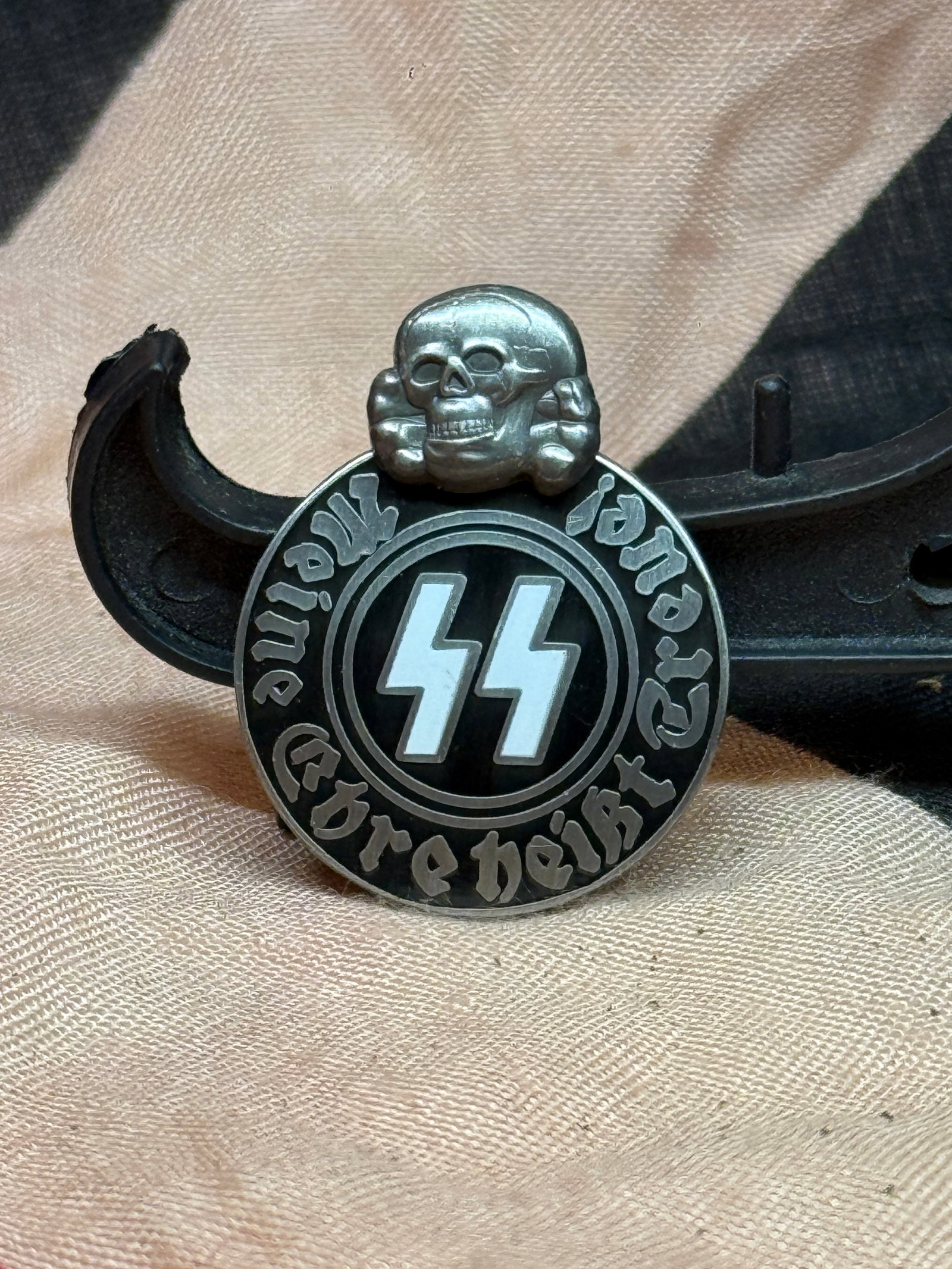 WWII German SS Motto Badge with Totenkopf, RZM M1/52 Marked: This circular metal badge displays the SS runes beneath a prominent Totenkopf (death’s head) motif and bears the inscription “Meine Ehre heißt Treue” (“My Honor is Loyalty”), the of