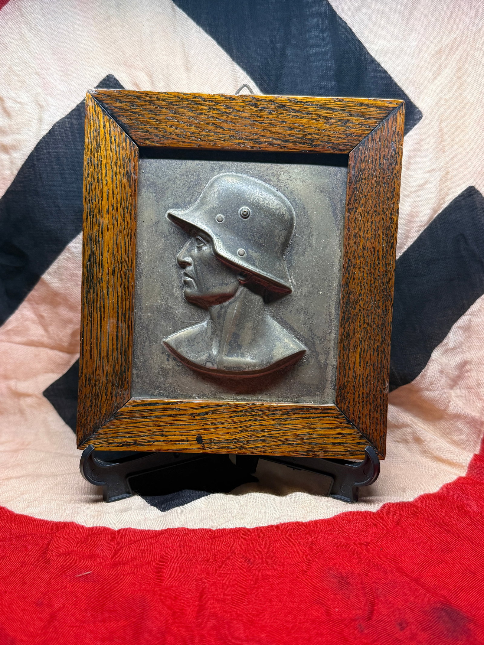 WWII German Soldier Relief Plaque in Wooden Frame: This framed metal relief plaque portrays the profile of a German soldier wearing a steel combat helmet characteristic of the Second World War era. Executed in raised relief and mounted within a staine