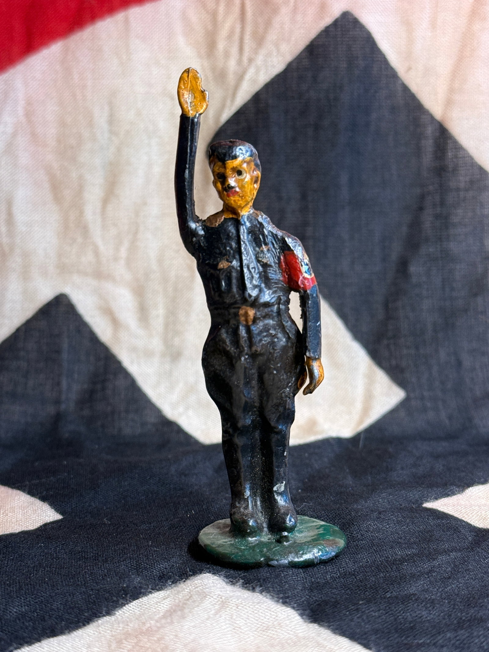 World War II German Hitler Youth Lead Toy Figure: Hand-painted cast-metal toy figure depicting a uniformed Hitler Youth member standing with raised arm in salute. The figure wears a dark uniform with a red armband and painted insignia details, mounte