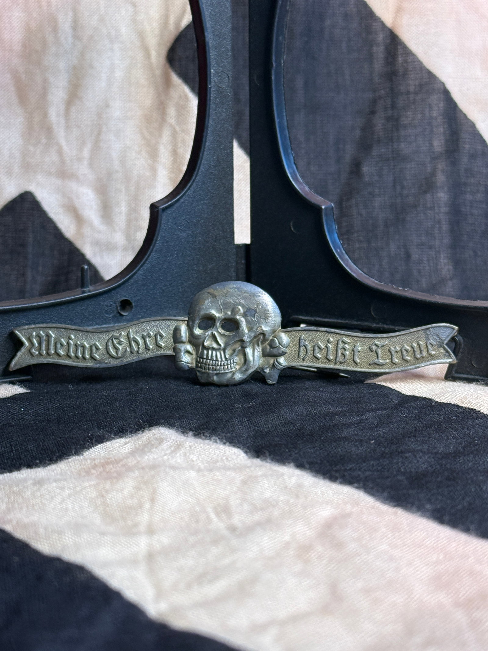 World War II German SS Totenkopf Evening Dress Insignia “Meine Ehre Heißt Treue: Metal SS evening dress insignia featuring a central Totenkopf (death’s head) skull and crossbones flanked by a scroll inscribed “Meine Ehre Heißt Treue” (“My Honor Is Loyalty”), the