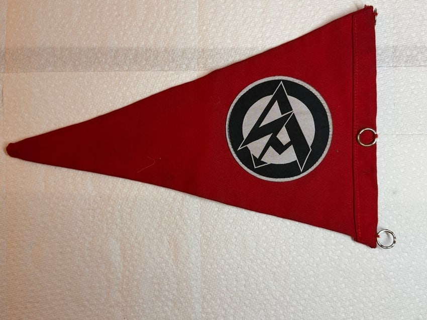 World War II German NSKK Organizational Pennant: Red triangular pennant displaying the emblem of the Nationalsozialistisches Kraftfahrkorps (NSKK), or National Socialist Motor Corps. Constructed of red cloth with a printed NSKK insignia enclosed wit