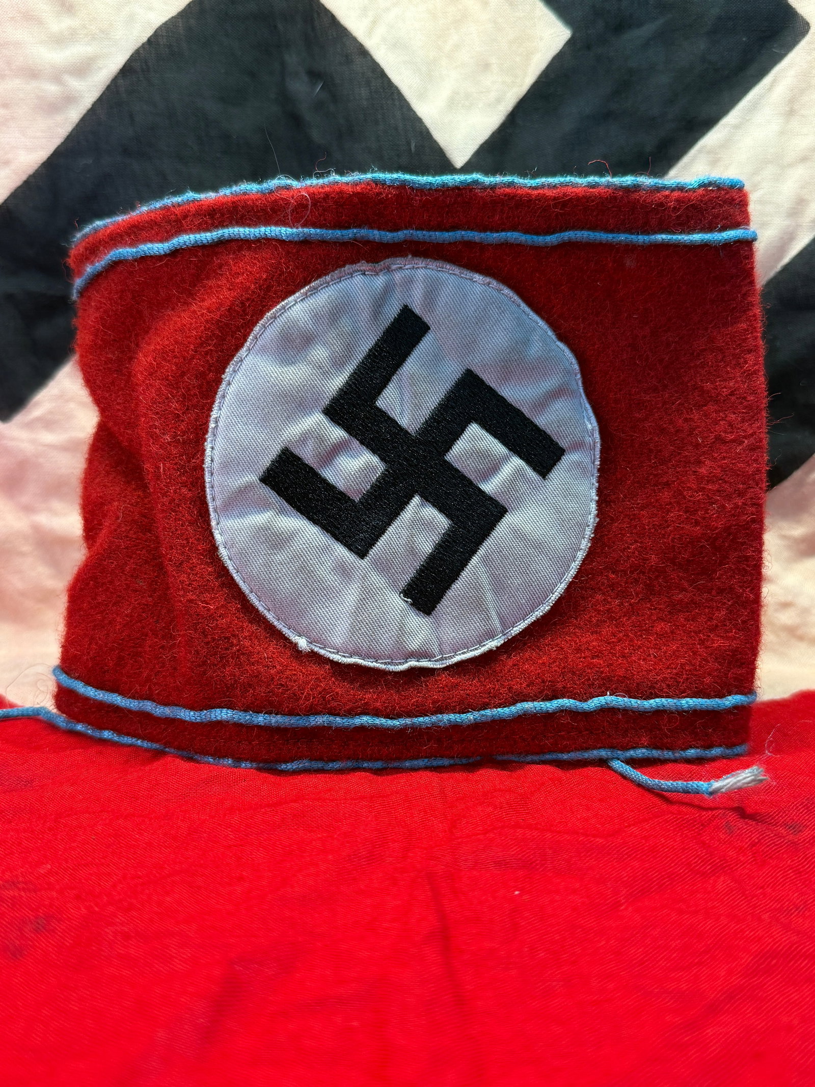 WWII German NSDAP Political Leader Armband: Red wool political leader armband trimmed with light blue piping and centered by a white roundel bearing a black swastika. The construction features stitched wool panels with blue cord edging and tie 