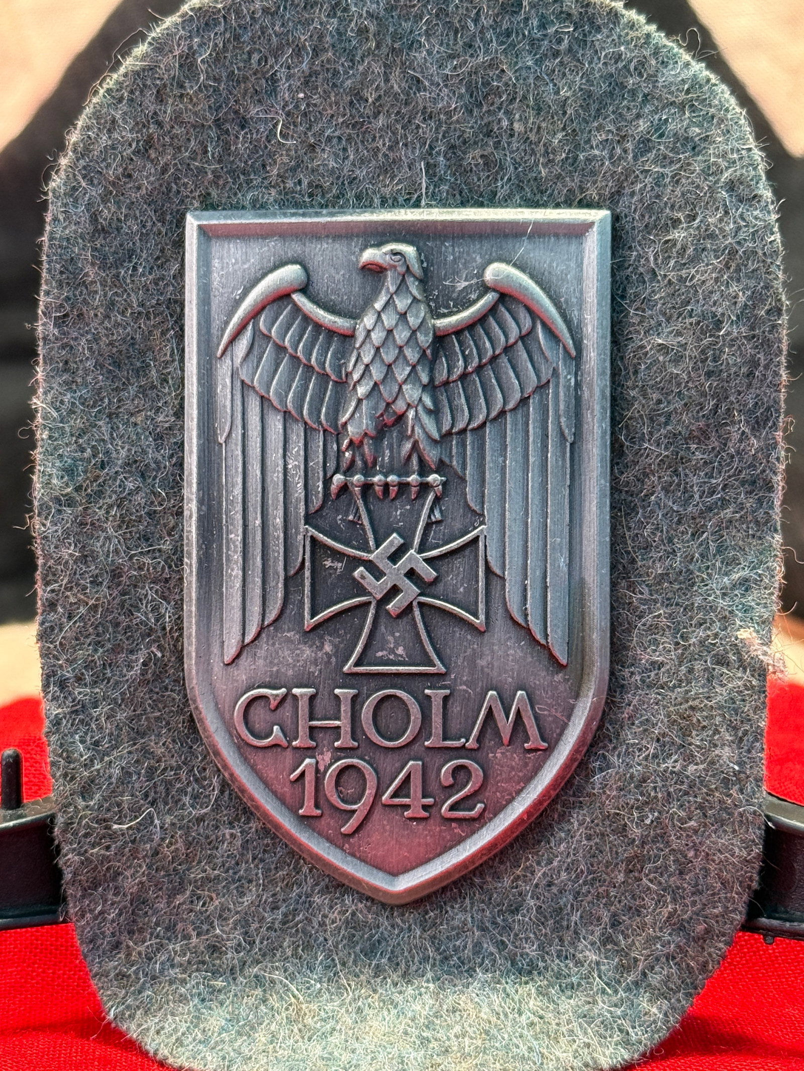 WWII German Cholm 1942 Campaign Shield (Cholmschild): Metal campaign shield depicting a German national eagle perched above an Iron Cross with the inscription "CHOLM 1942." The reverse retains its original attachment plate with prongs secured through a g