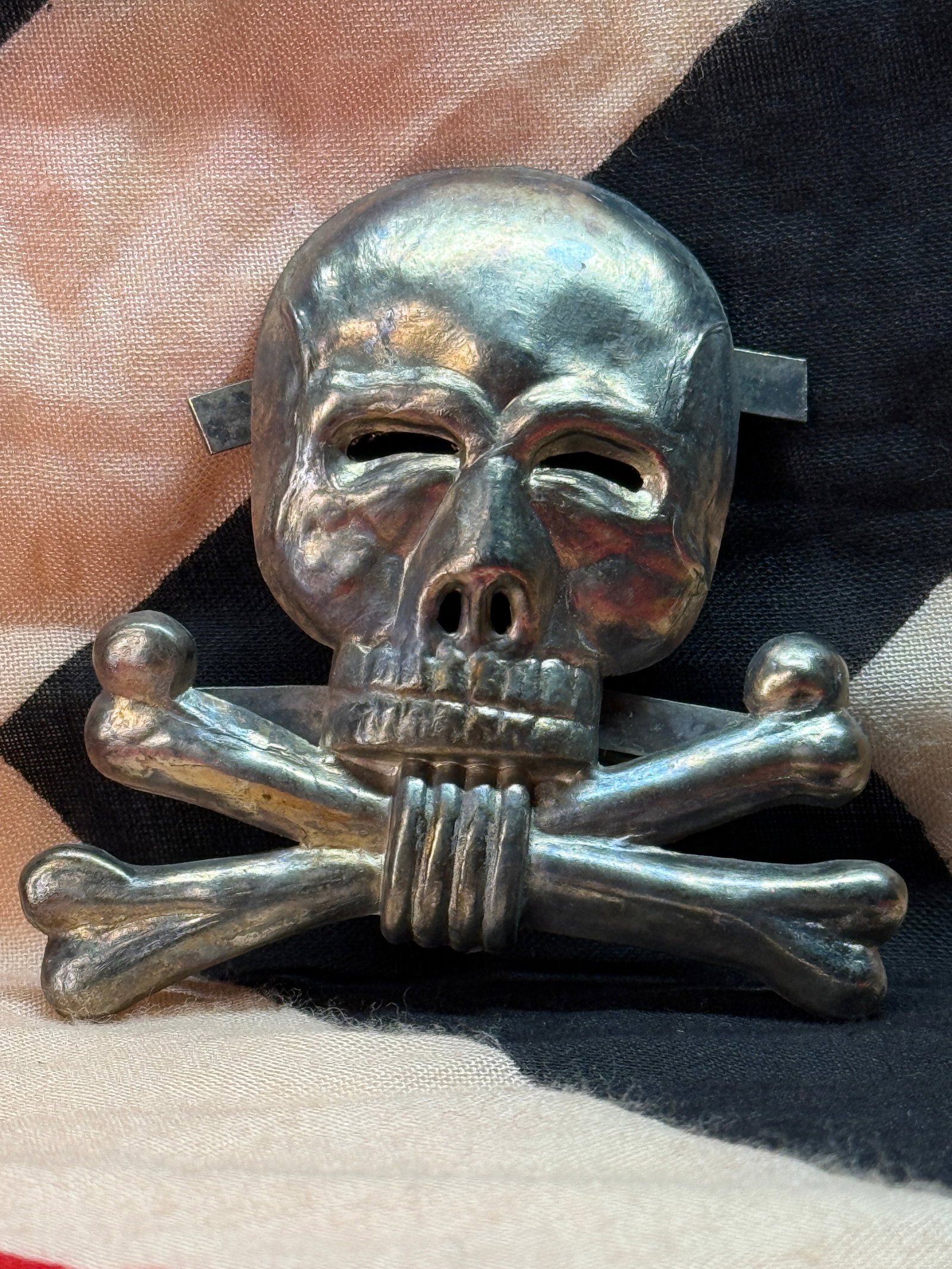 WWII German SS Totenkopf Cap Insignia: This three-dimensional metal Totenkopf (Death's Head) insignia features a detailed skull above crossed bones with rear attachment tabs for mounting to military headgear. The Totenkopf served as one of