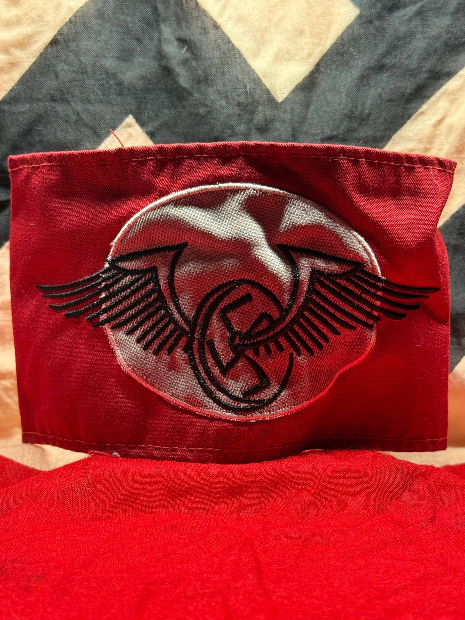 WWII German National Socialist Flyers Corps (NSFK) Armband: This red cloth armband features a white roundel embroidered with the winged emblem of the National Socialist Flyers Corps (NSFK) incorporating a central swastika. The reverse reveals period-style sewn