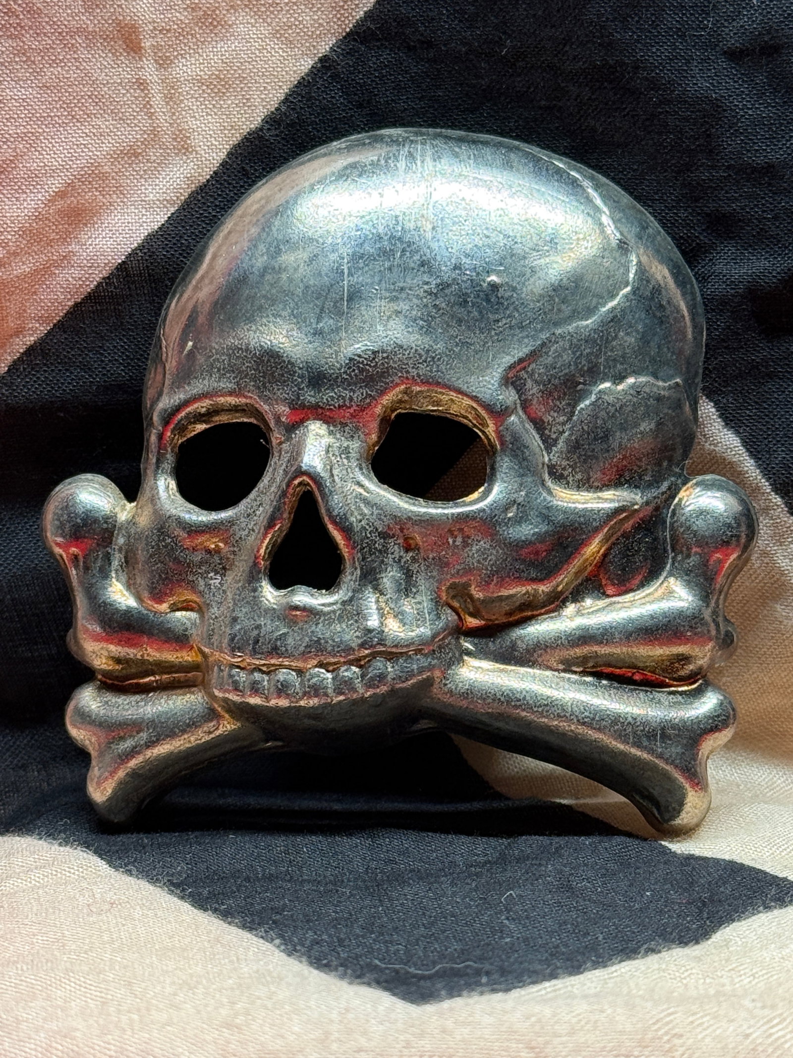 WWII German SS Totenkopf Cap Badge: This WWII German SS Totenkopf cap badge features a skull-and-crossbones design with hollow eye sockets and rear attachment prongs. The metal surface shows age, patina, and wear, with a bold three-dime