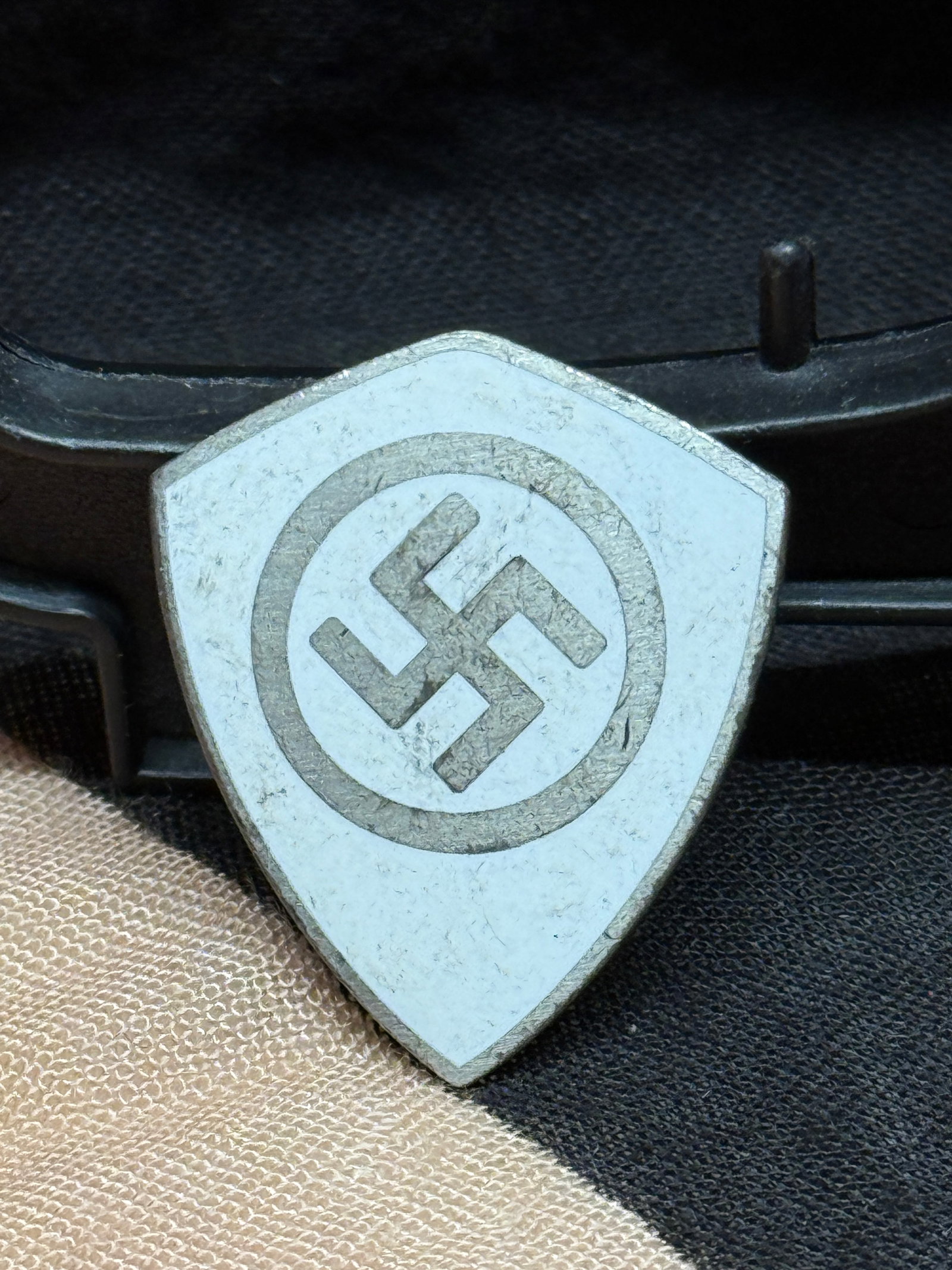 WWII German Reich Labor Service (RAD) Enamel Membership Badge: This shield-shaped badge features a black swastika within a circular border set against a white enamel field. The reverse retains its original pinback assembly and is marked "GES. GESCH." indicating l