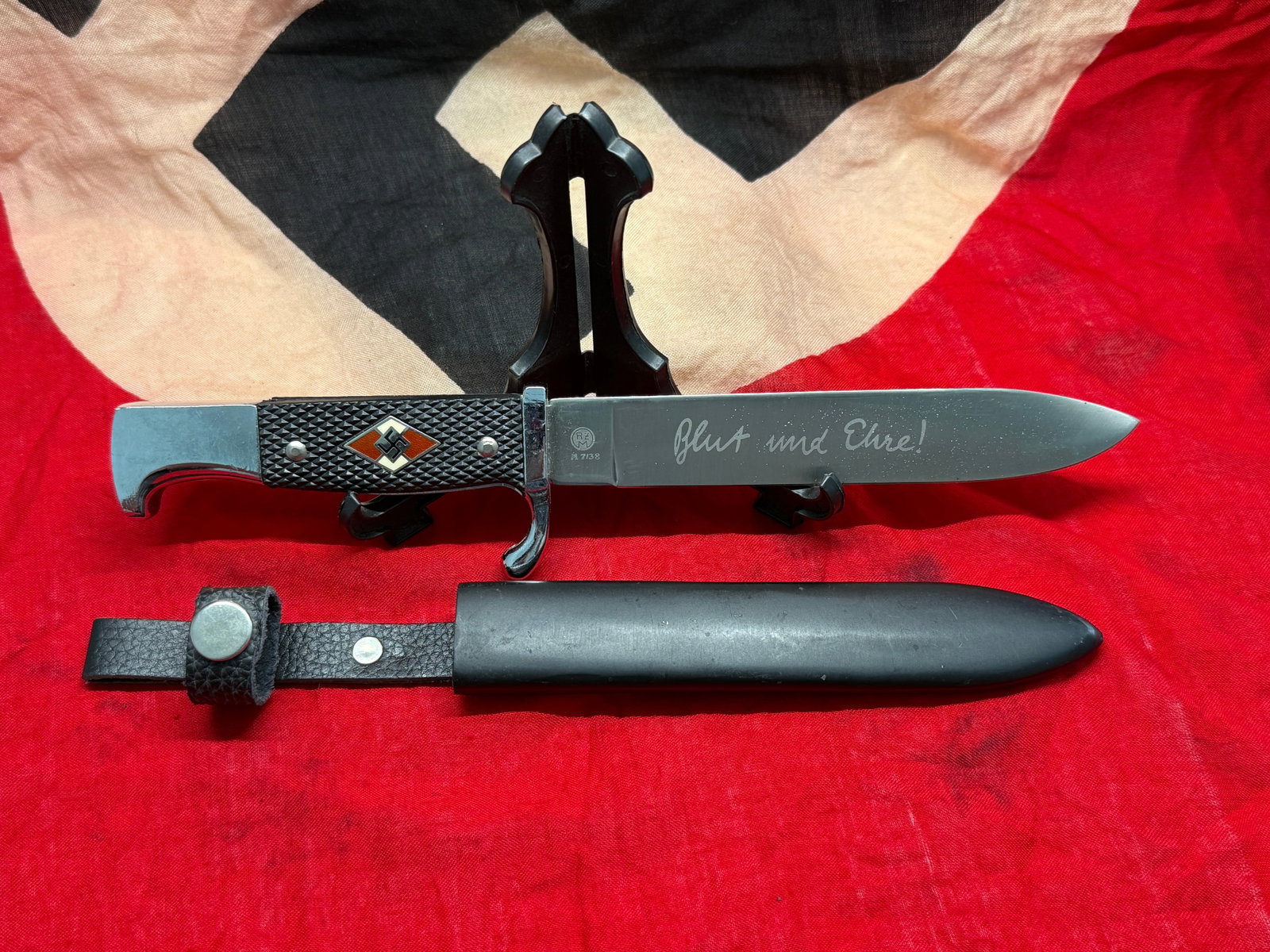 Hitler Youth (HJ) Utility Knife with Scabbard: This fixed-blade utility knife features the distinctive black checkered grip inset with the Hitler Youth diamond emblem, a plated guard and pommel, and a black metal scabbard with its leather belt han
