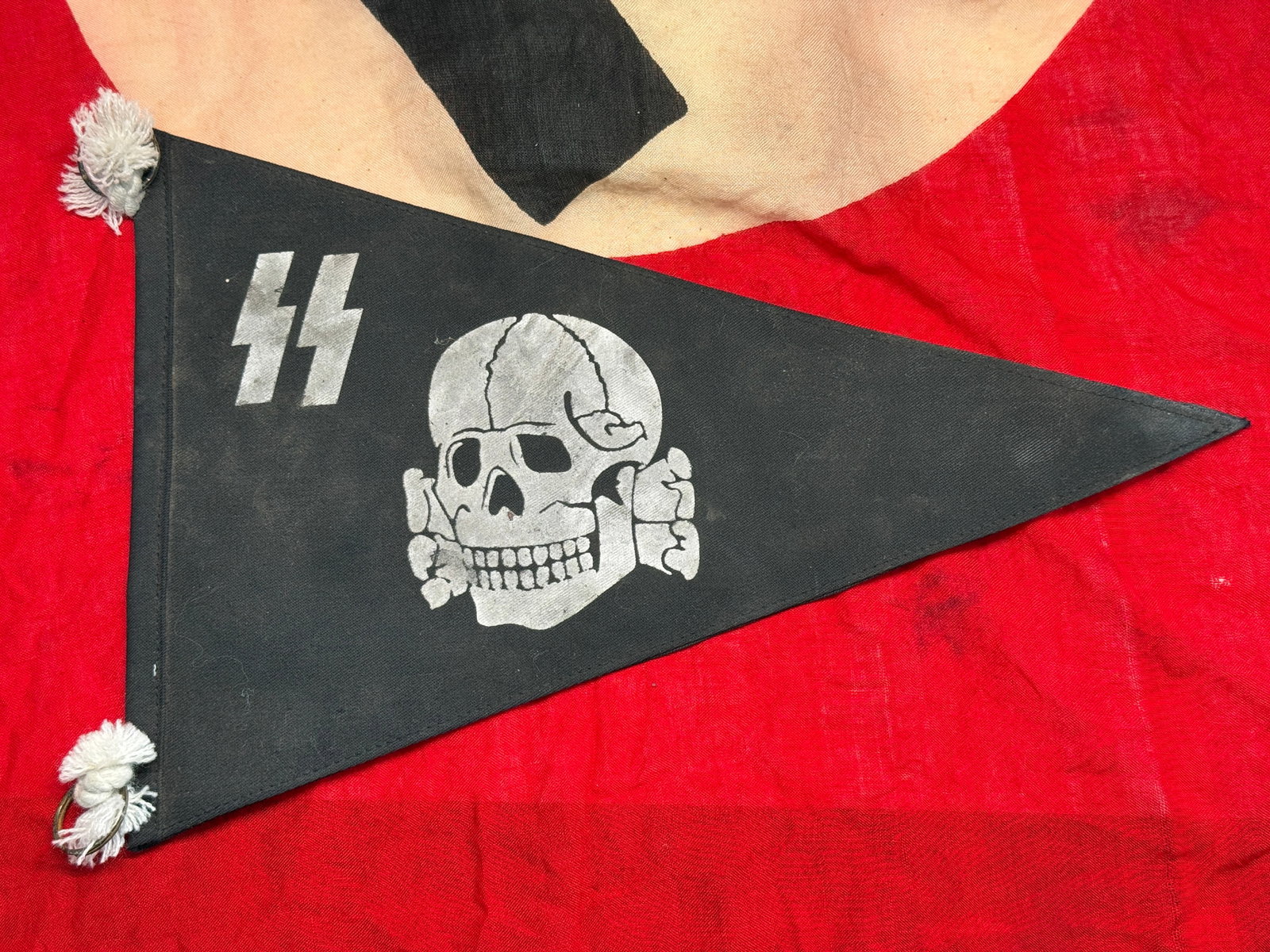 WWII German Waffen-SS Totenkopf Pennant: This triangular black cloth pennant displays the iconic Waffen-SS Totenkopf (Death's Head) emblem accompanied by the SS runes, with white tassels and suspension rings at the hoist. Pennants of this st