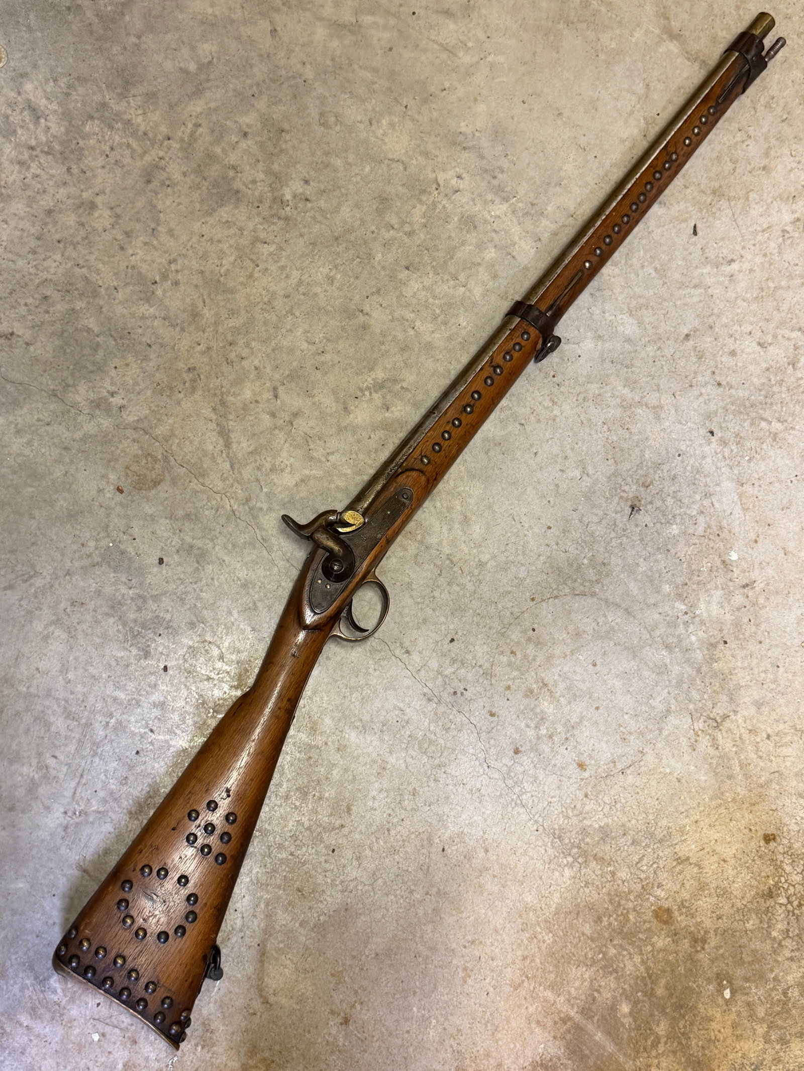 Indian Wars 1870s Native American Indian Studded Flintlock Musket: This 1870s Native American Indian Studded Flintlock Musket features a full-length hardwood stock decorated with extensive brass tackwork forming distinctive geometric patterns across the buttstock and
