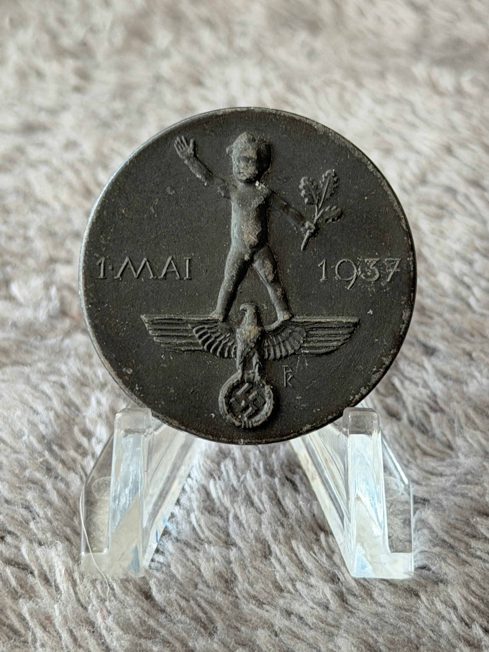 WWII German 1937 May Day (1. Mai) Tinnie Badge – H. Hansenmayer Pforzheim: This WWII German 1937 May Day (1. Mai) Tinnie Badge features a child standing atop a Third Reich eagle and wreath, raising one hand in salute while holding an oak branch, with the date "1. Mai 1937" p